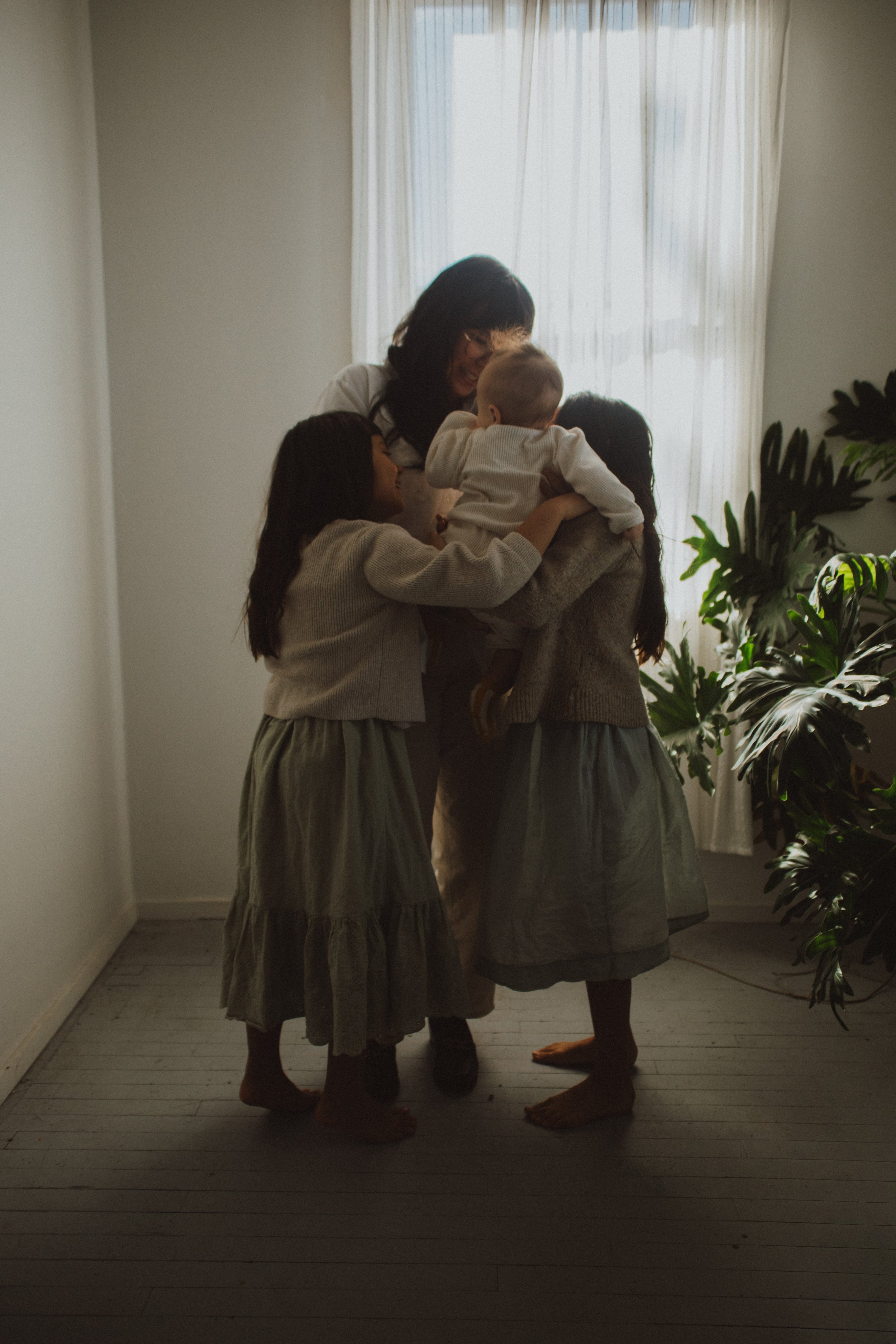 A woman and three girls hugging and holding a small child in a room with a window and green plants.