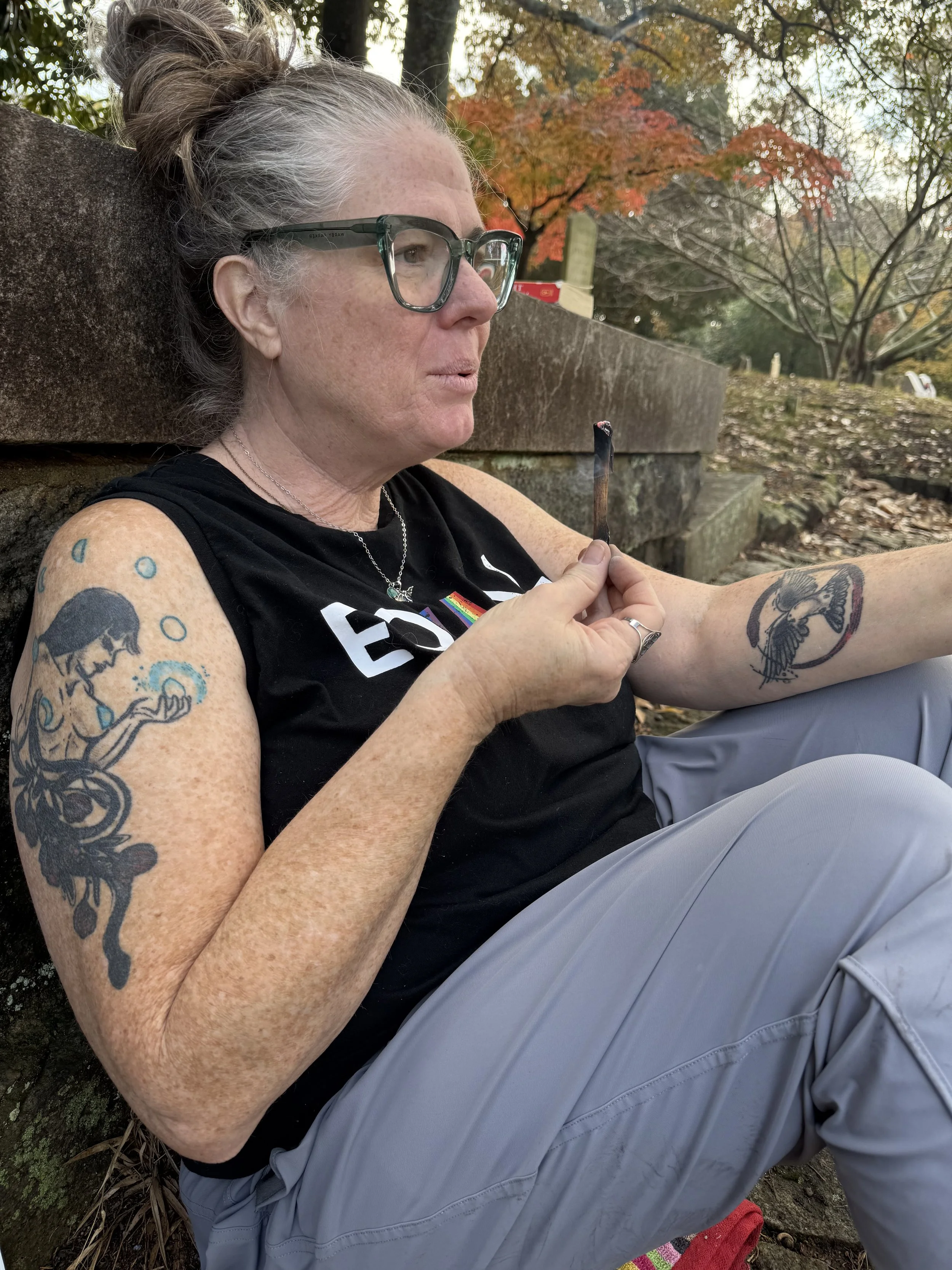 A woman with tattoos and glasses sitting outdoors in fall, smoking a rolled cigarette or joint.