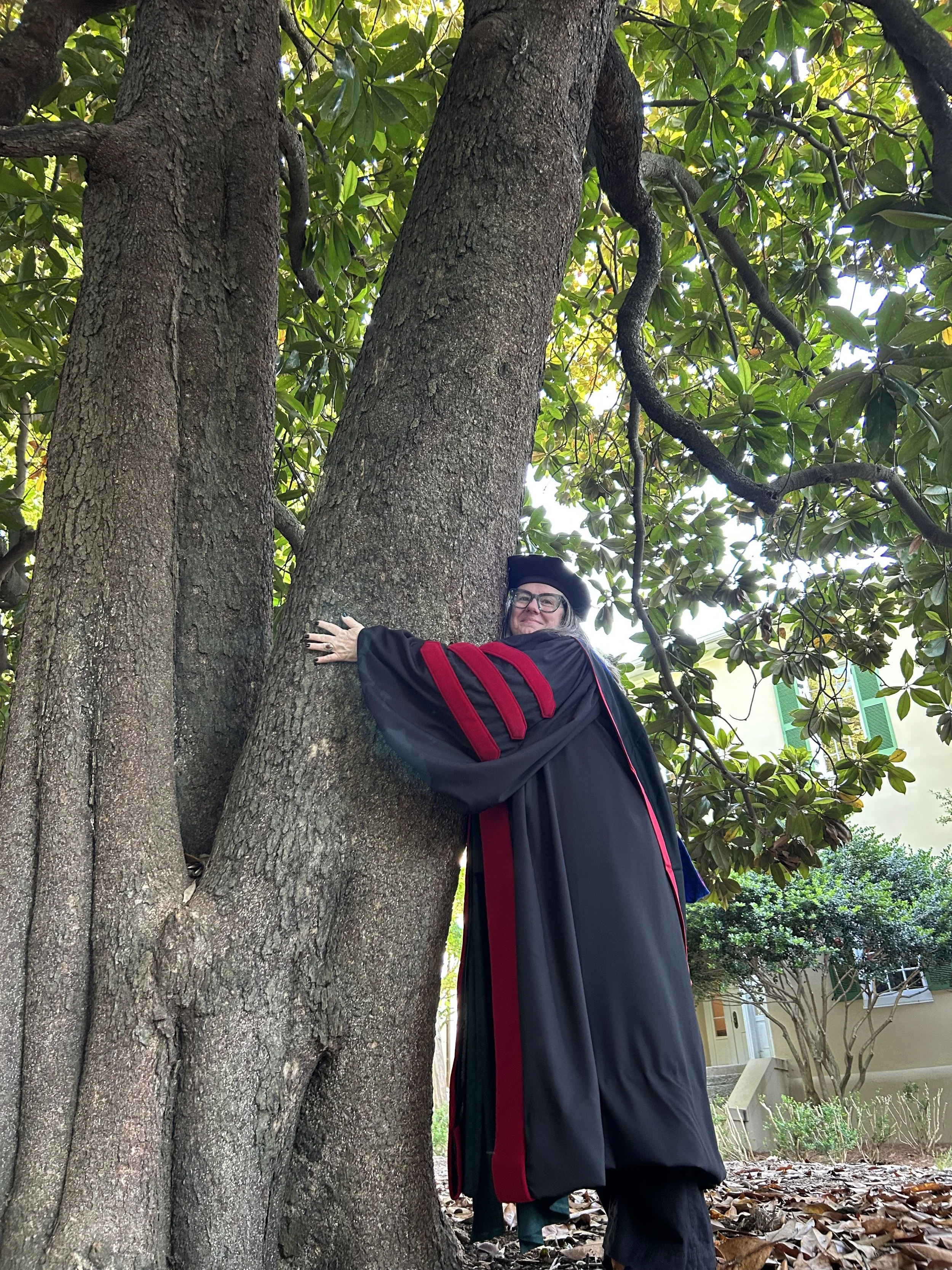 A person wearing academic regalia hugging a large tree outdoors surrounded by leaves and greenery.
