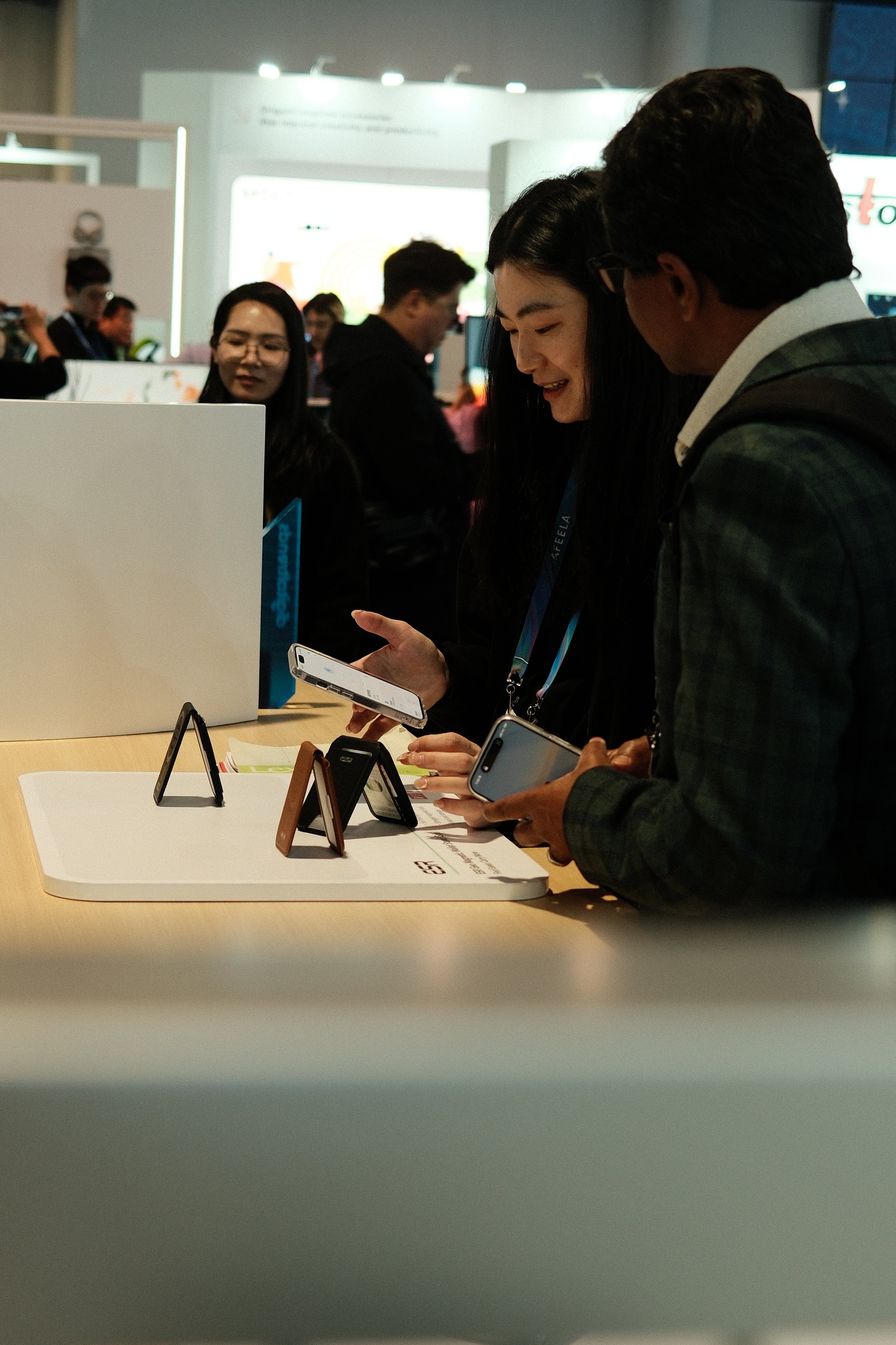 Two people are looking at smartphones displayed on a table at a tech event, with several other attendees in the background.