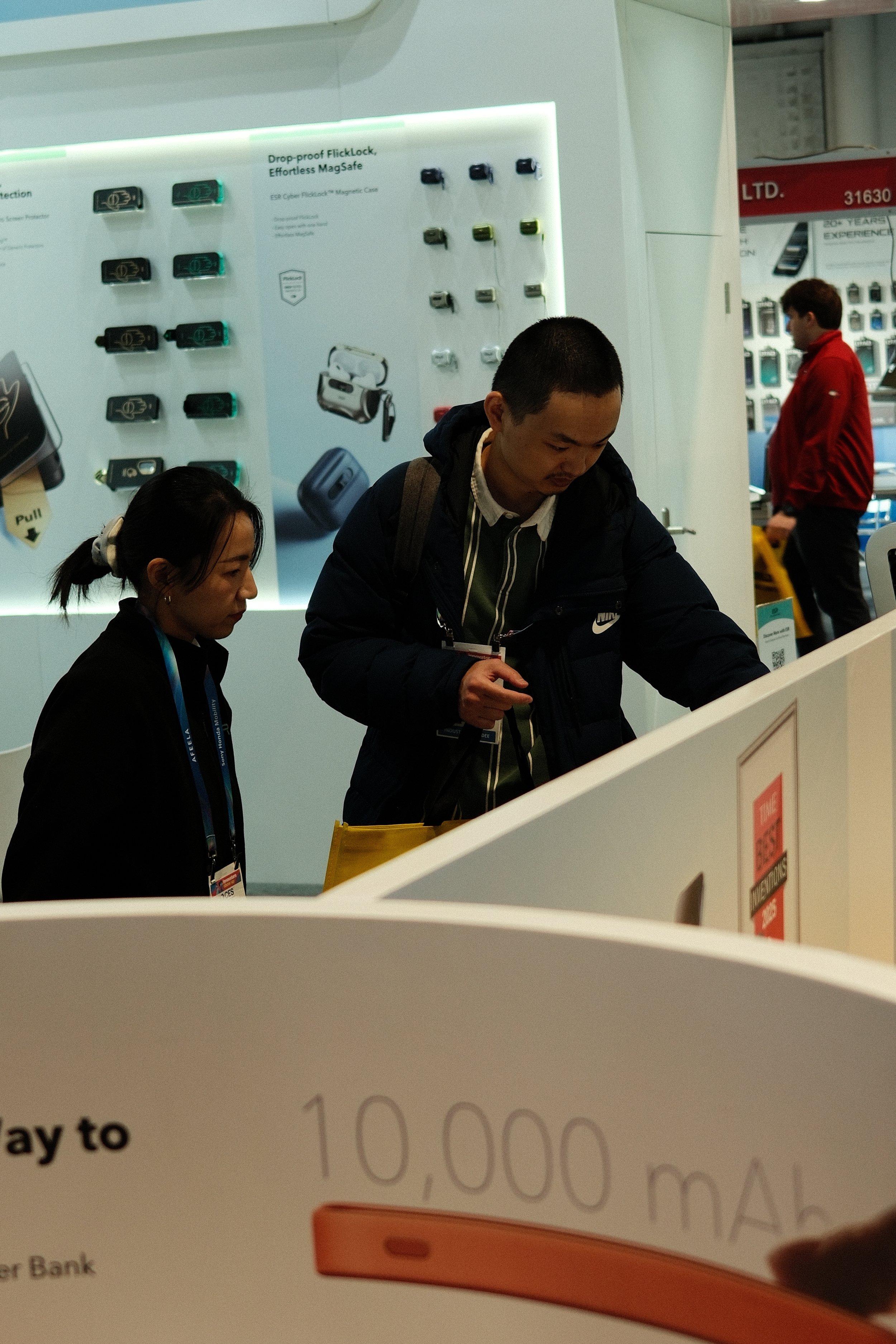 Two people are standing at a counter or display, looking at something. In the background, there are display panels with various electronic accessories, including cases and chargers.