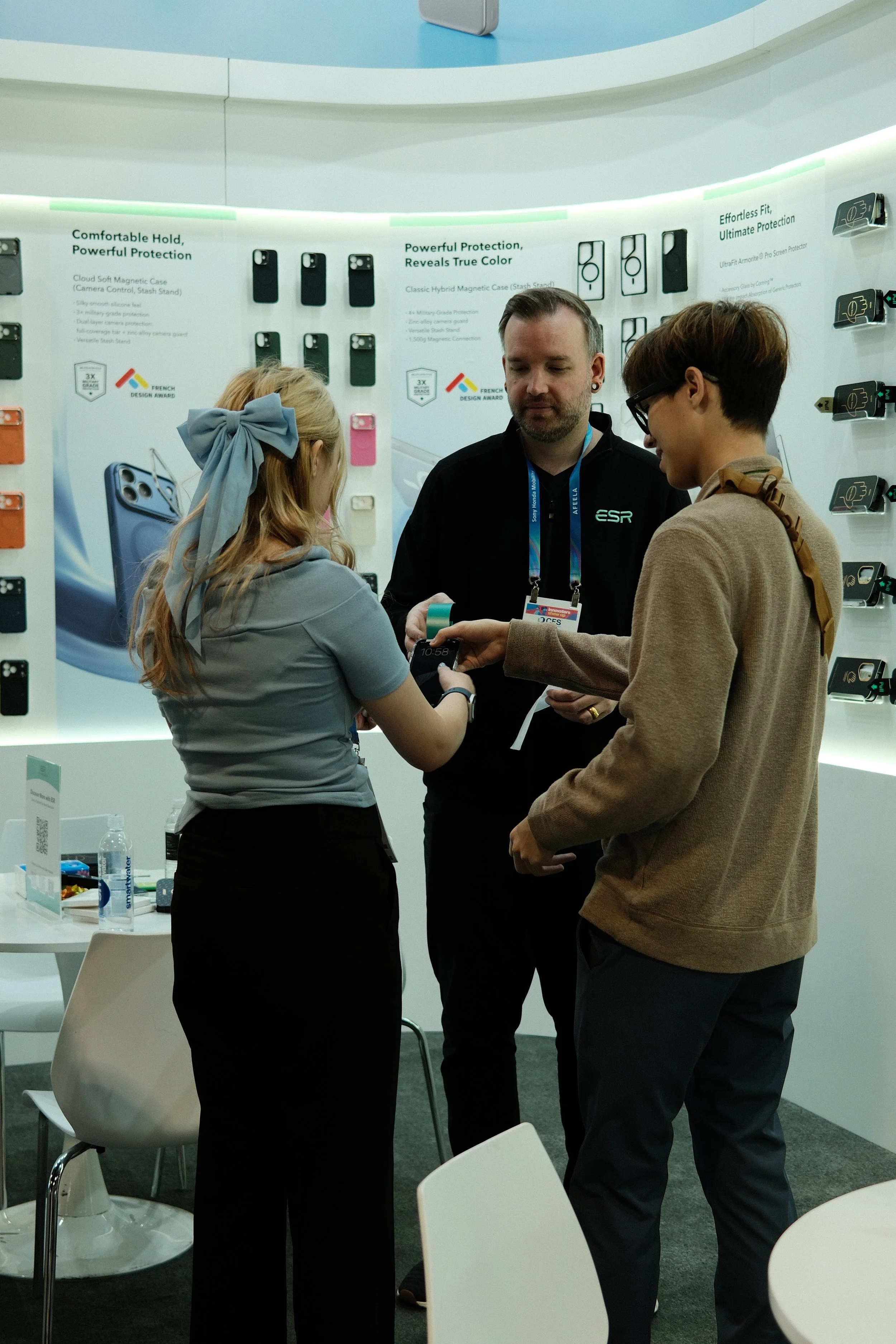 Three people are standing at a booth displaying various smartphone cases, with one person showing something on their smartwatch to the others.