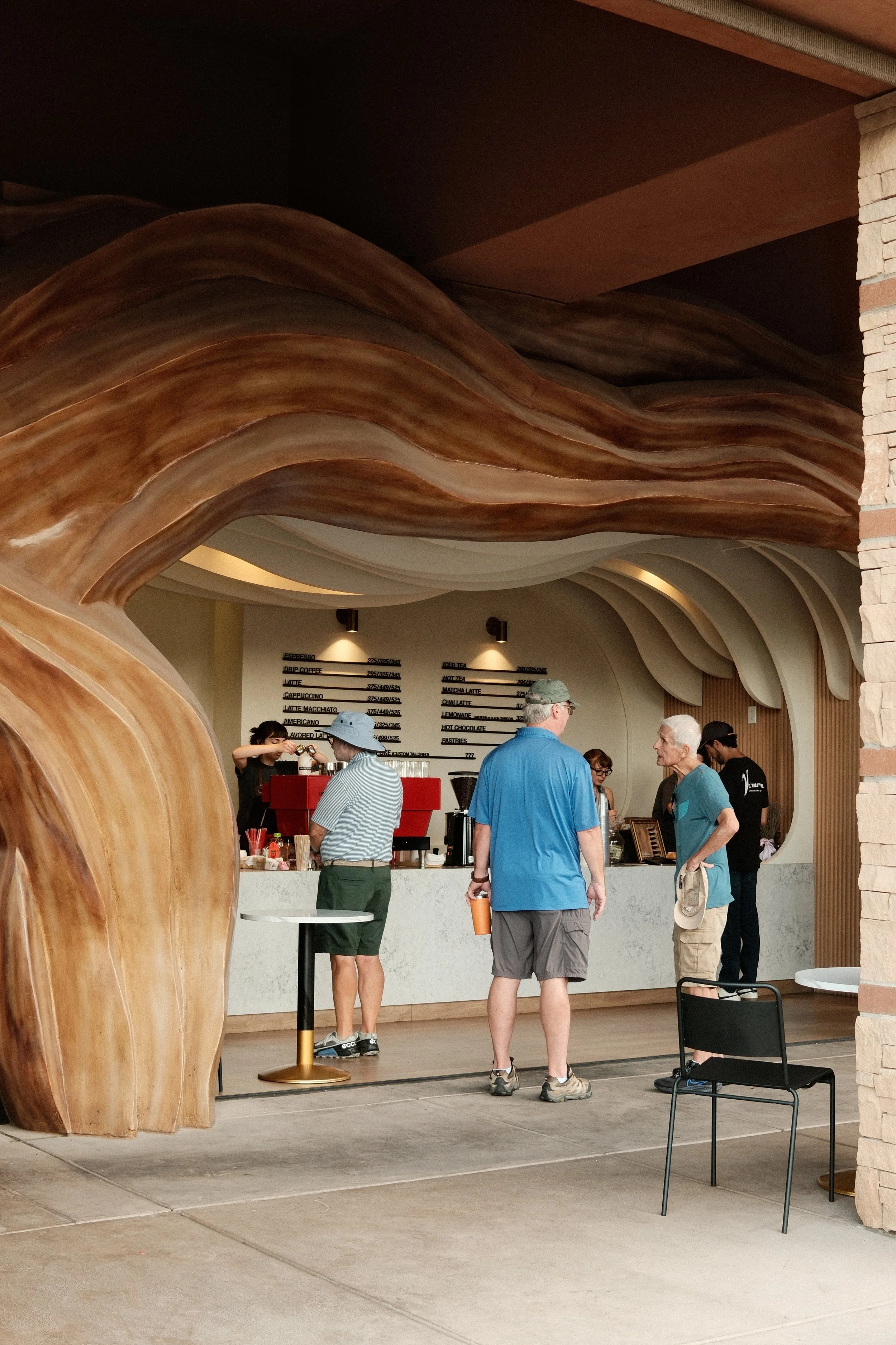 People ordering coffee at a cafe counter inside a building with a modern, artistic interior design featuring large, curved wooden structures and a white, abstract ceiling.