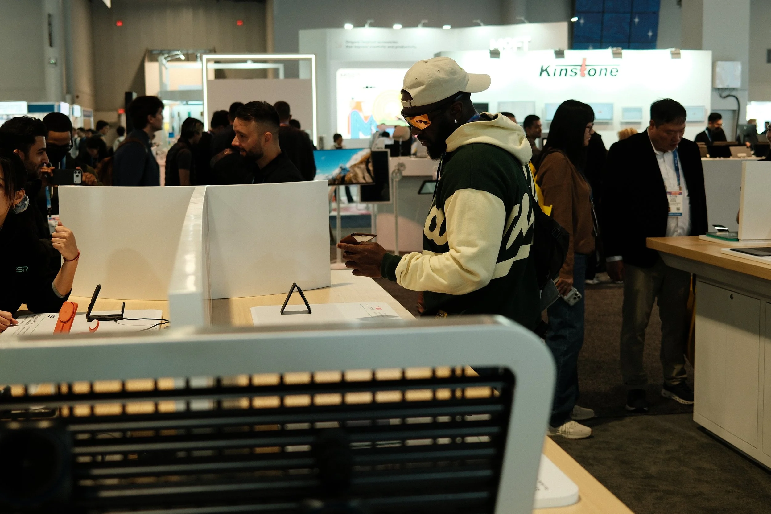 A crowded technology trade show with people interacting at various booths, including a man wearing a hoodie and cap looking at his phone.