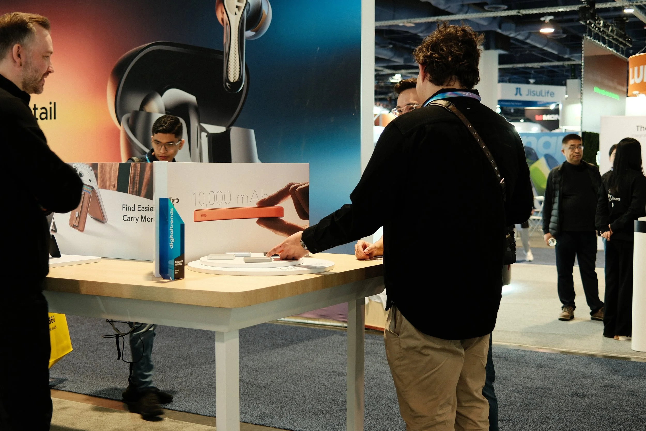 People browsing a display at a technology trade show, with posters promoting a wireless earbuds case and a 10,000 mAh power bank.