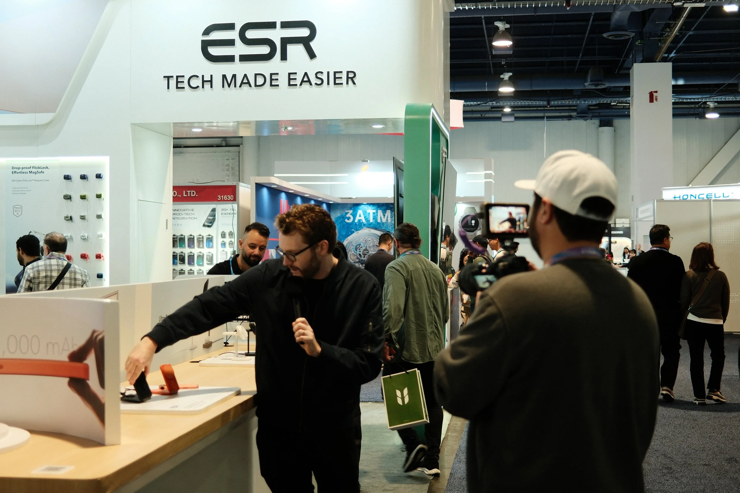 People attending a technology trade show booth with the ESG logo and the slogan 'Tech Made Easier'. A man is filming with a camera while another man displays a product on a table.