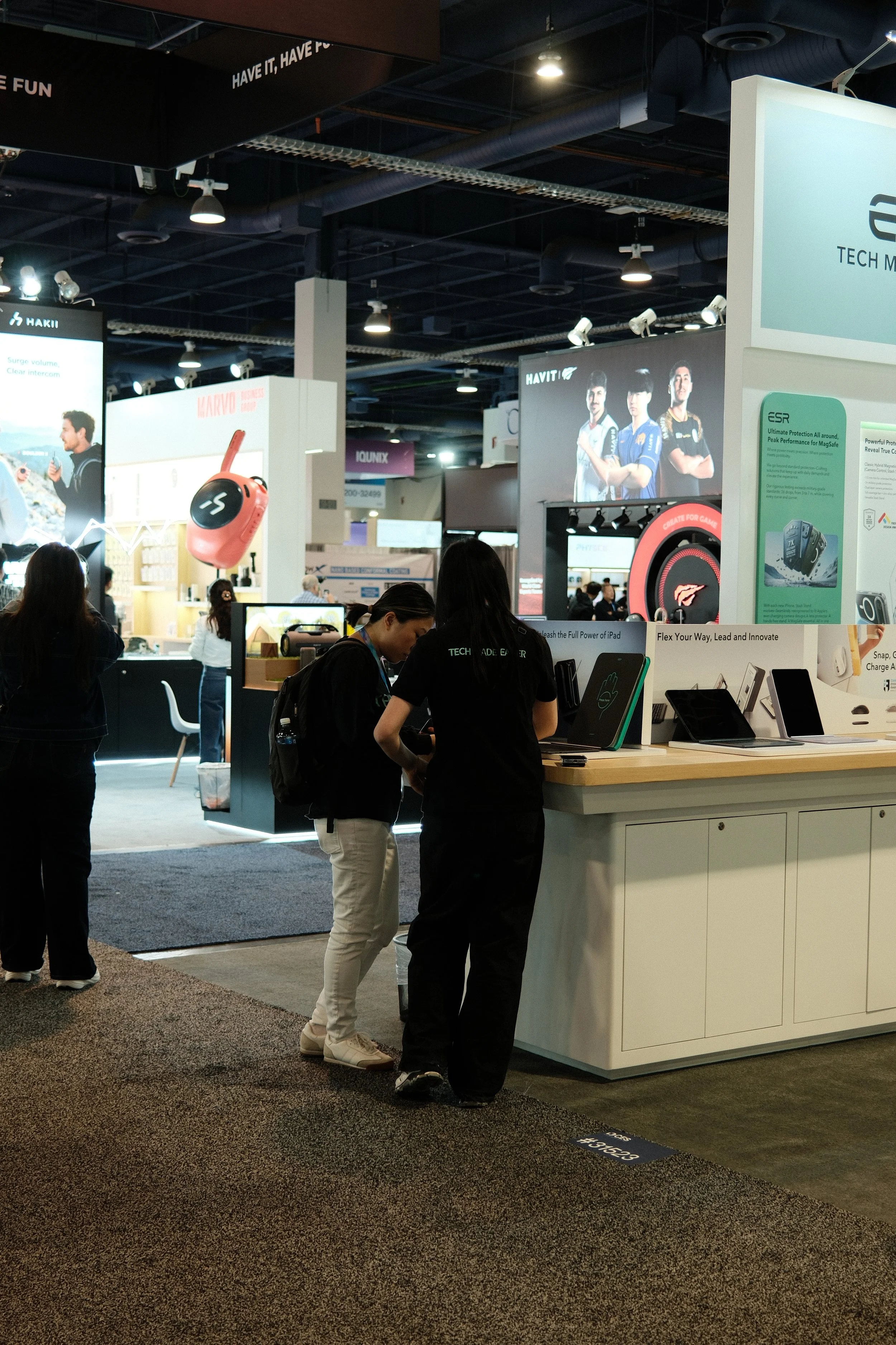 Two women in black shirts with 'Tech Made Easy' printed on the back are standing at a booth at a trade show, looking at tablets or electronic devices on display. Other attendees are seen in the background, with various exhibition booths and large scr