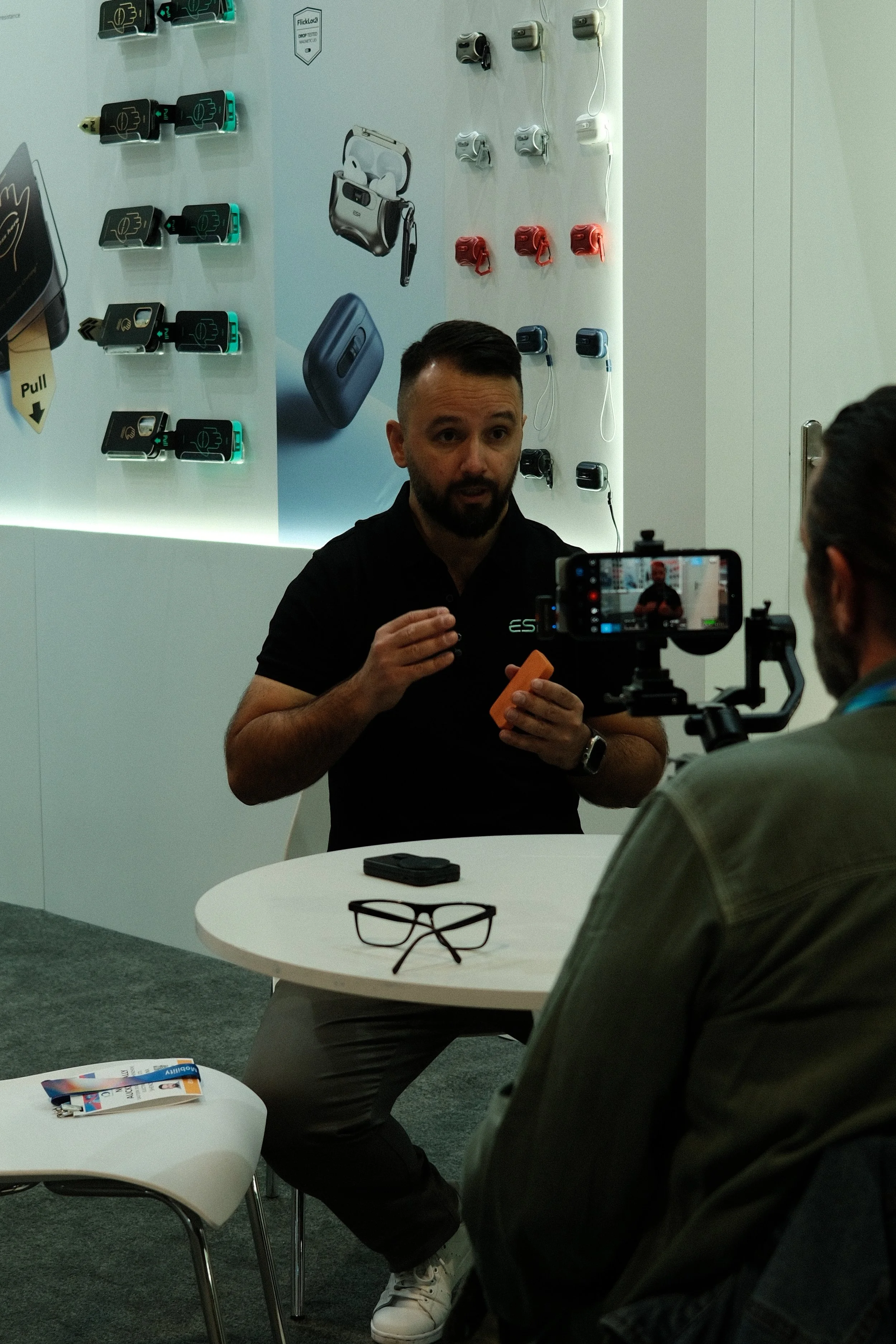 Man being interviewed at a display of electronic gadgets on a wall, including earphones, portable speakers, and wireless earbuds.