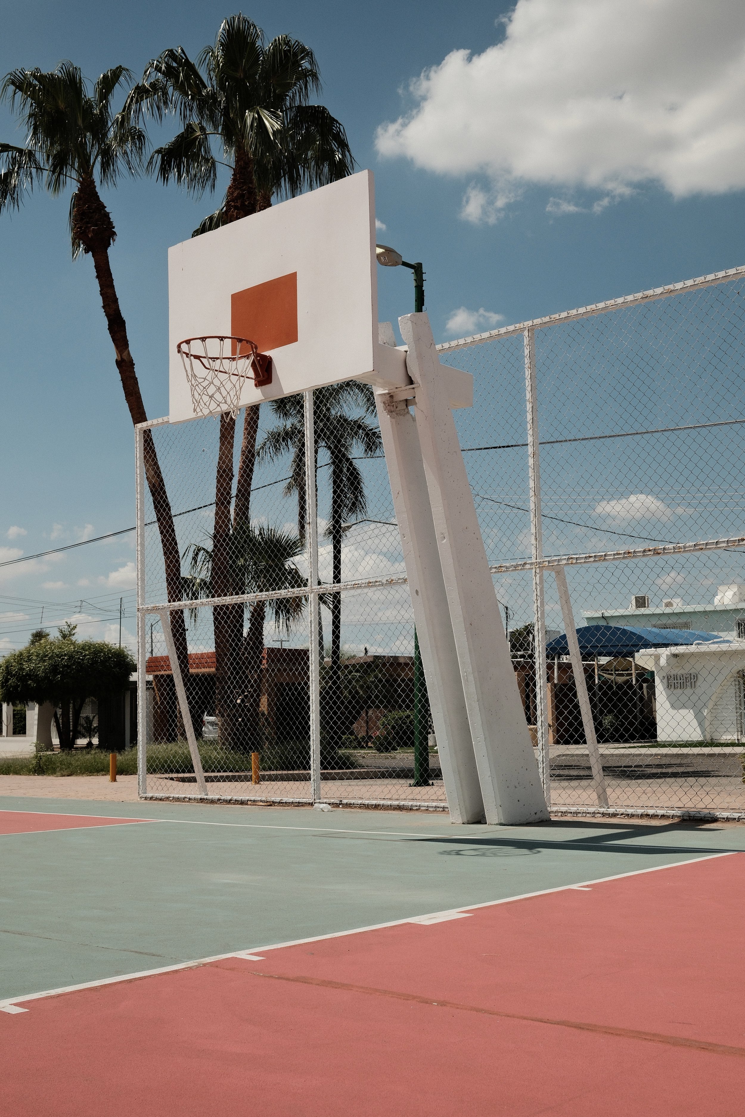 An outdoor basketball court with a damaged basketball hoop and backboard, palm trees, and a partly cloudy sky.