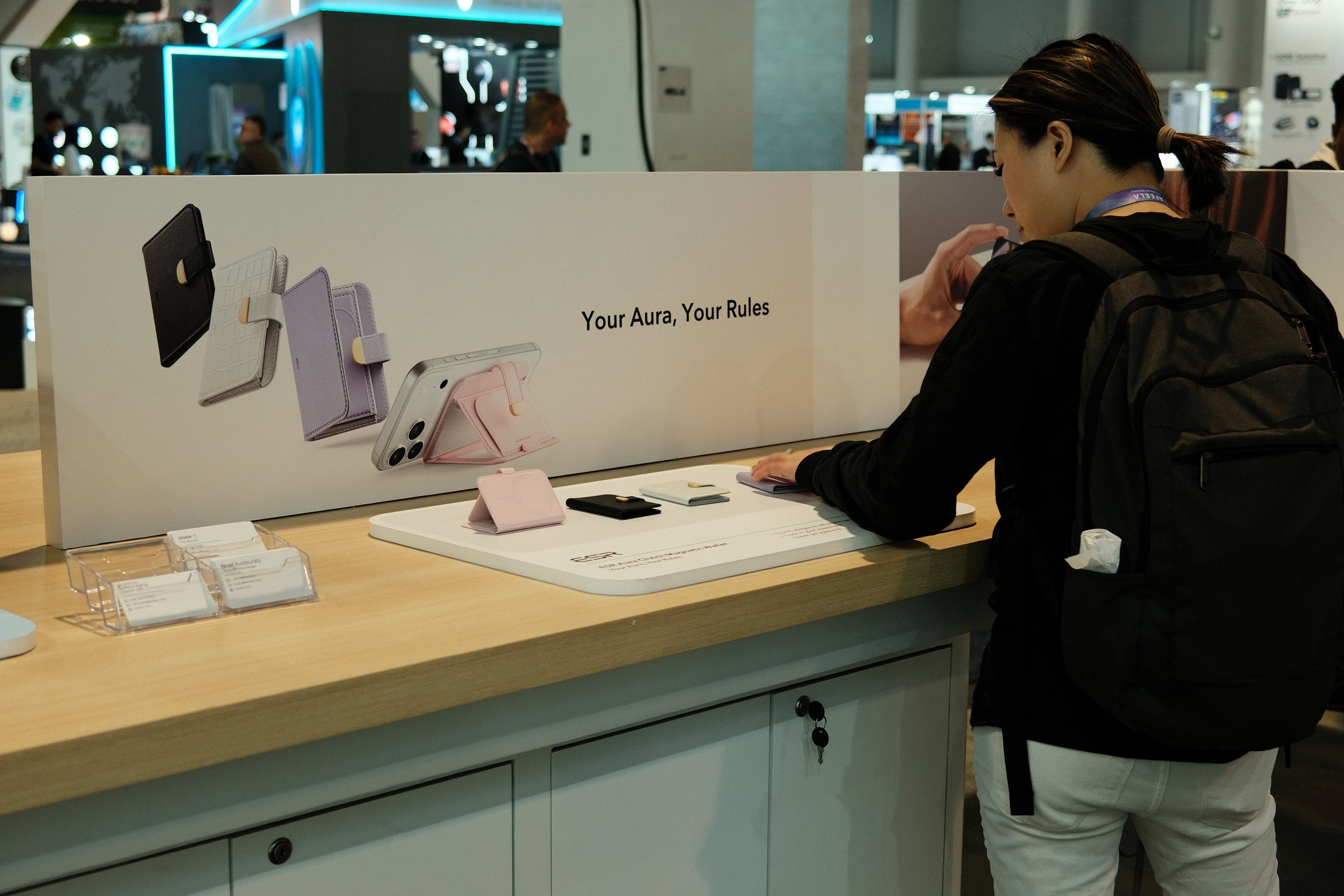 A woman with a ponytail and black backpack viewing phone accessories at a display table in an indoor shopping mall or exhibition center.