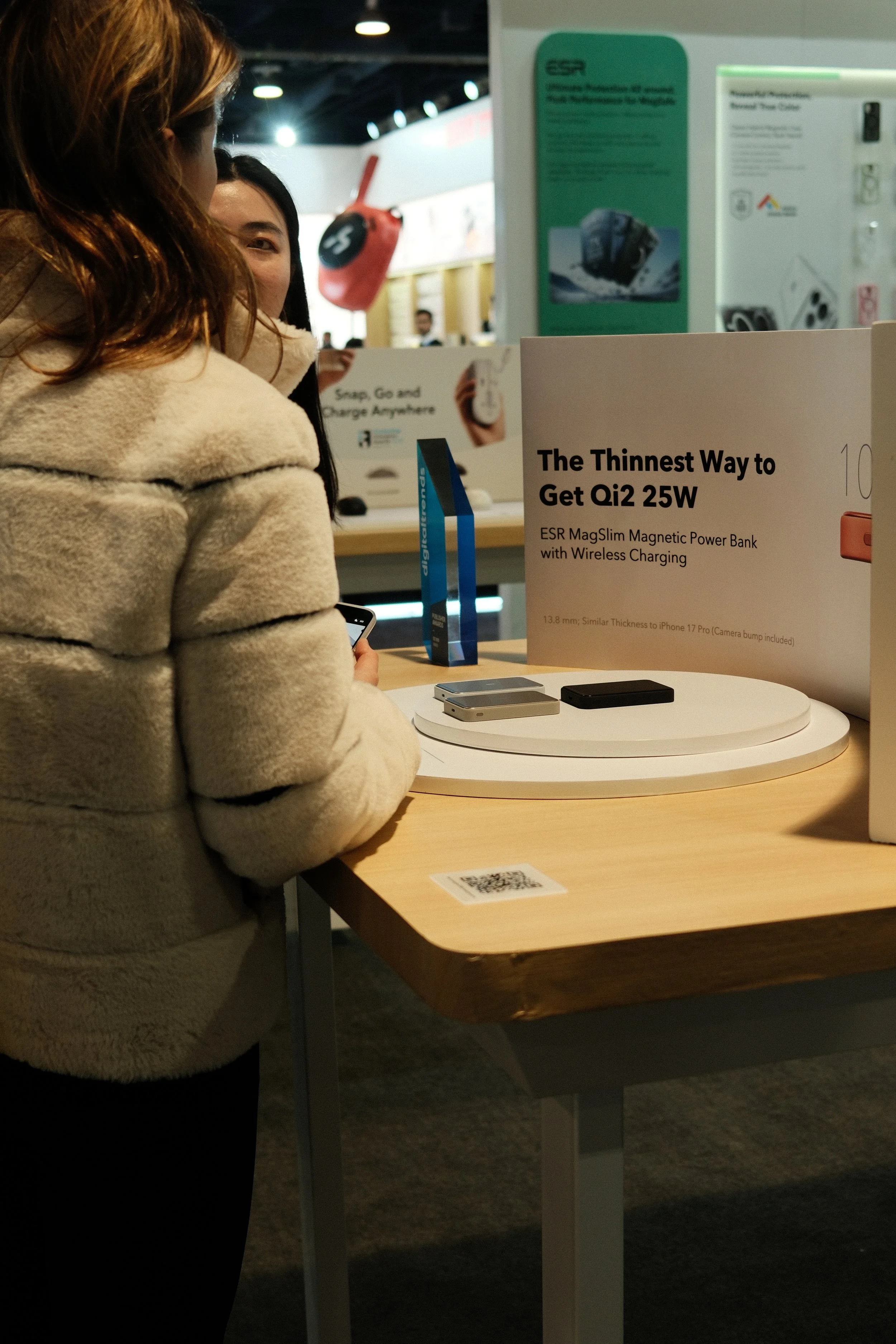 Two women standing at a display table with electronic devices in a store, with promotional signage about a magnetic power bank, and a QR code on the table.
