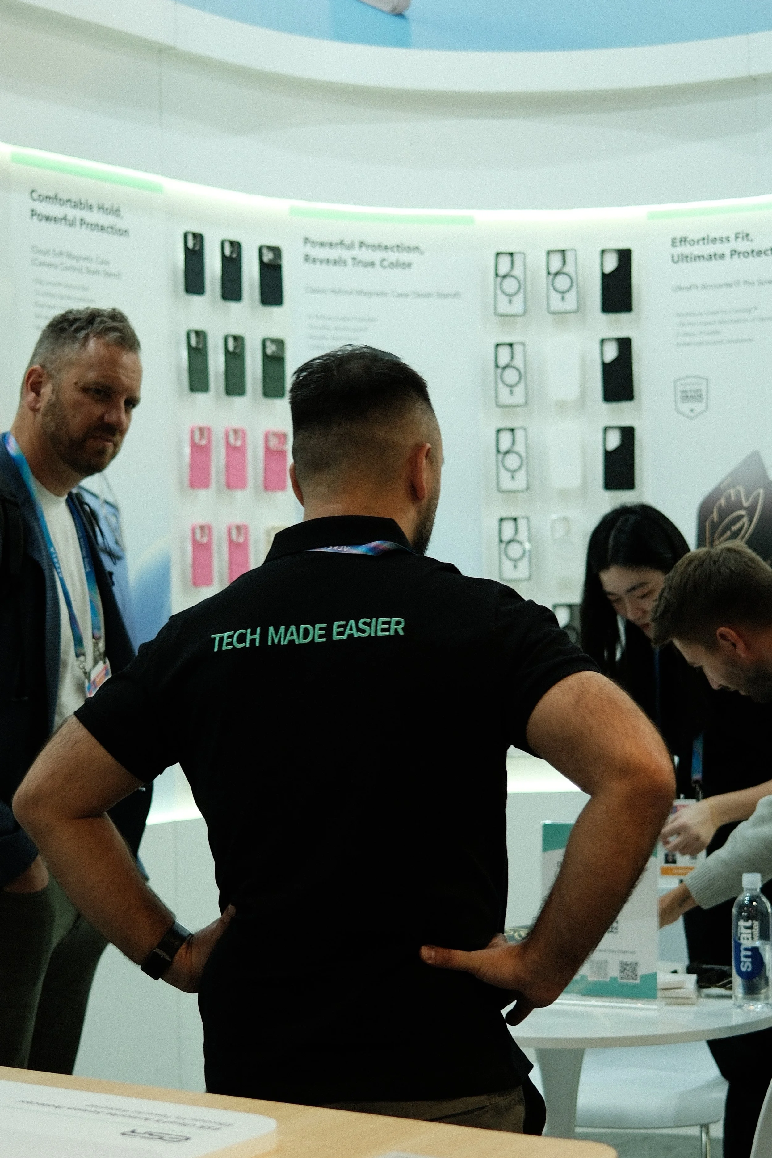 People at a technology trade show booth discussing products. One person wears a black shirt with "TECH MADE EASIER" printed on back. The booth has product displays and informational posters.