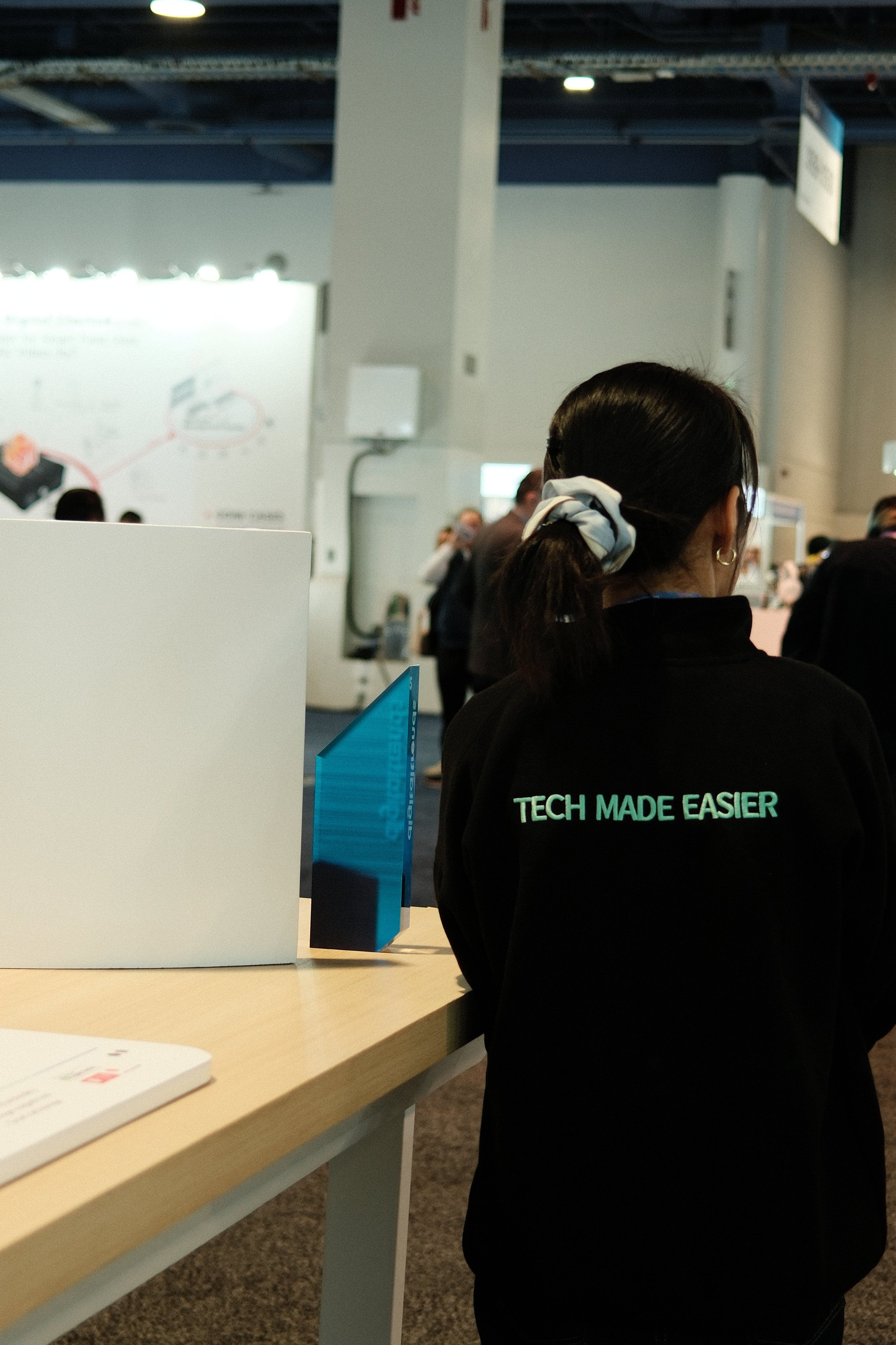 A woman with dark hair tied in a ponytail, wearing glasses and a black jacket with the words 'TECH MADE EASIER' on the back, standing at a booth in a busy indoor event or convention.