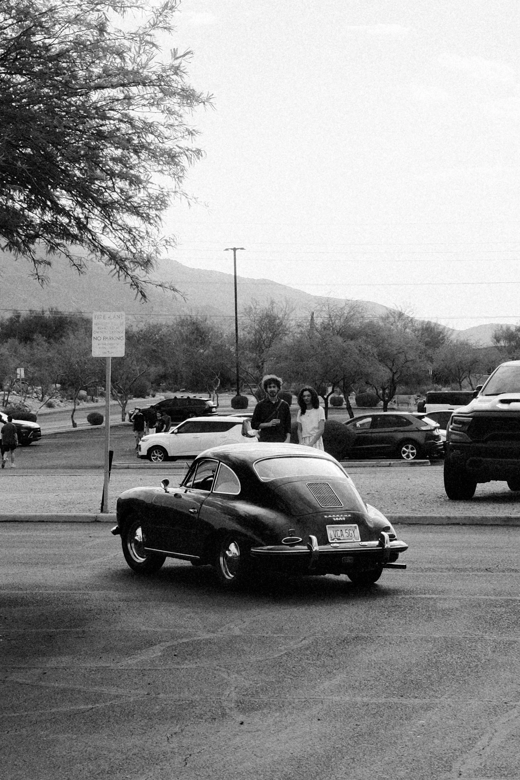 A black vintage car parked in a parking lot with two people standing behind it, surrounded by other cars, trees, mountains, and a sign in the background.