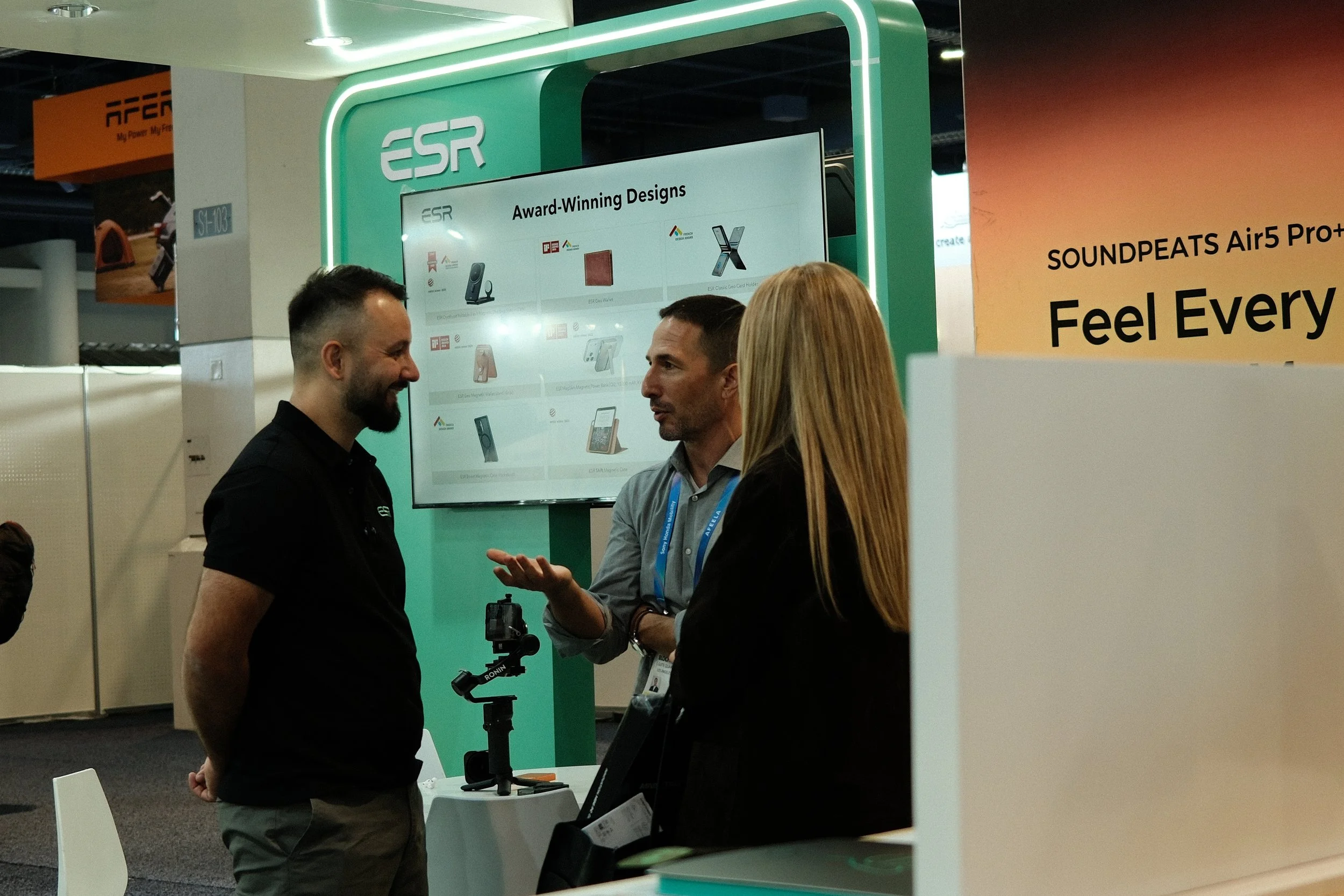 Three people engaged in a conversation at a trade show booth, with a display board showing award-winning designs in the background.