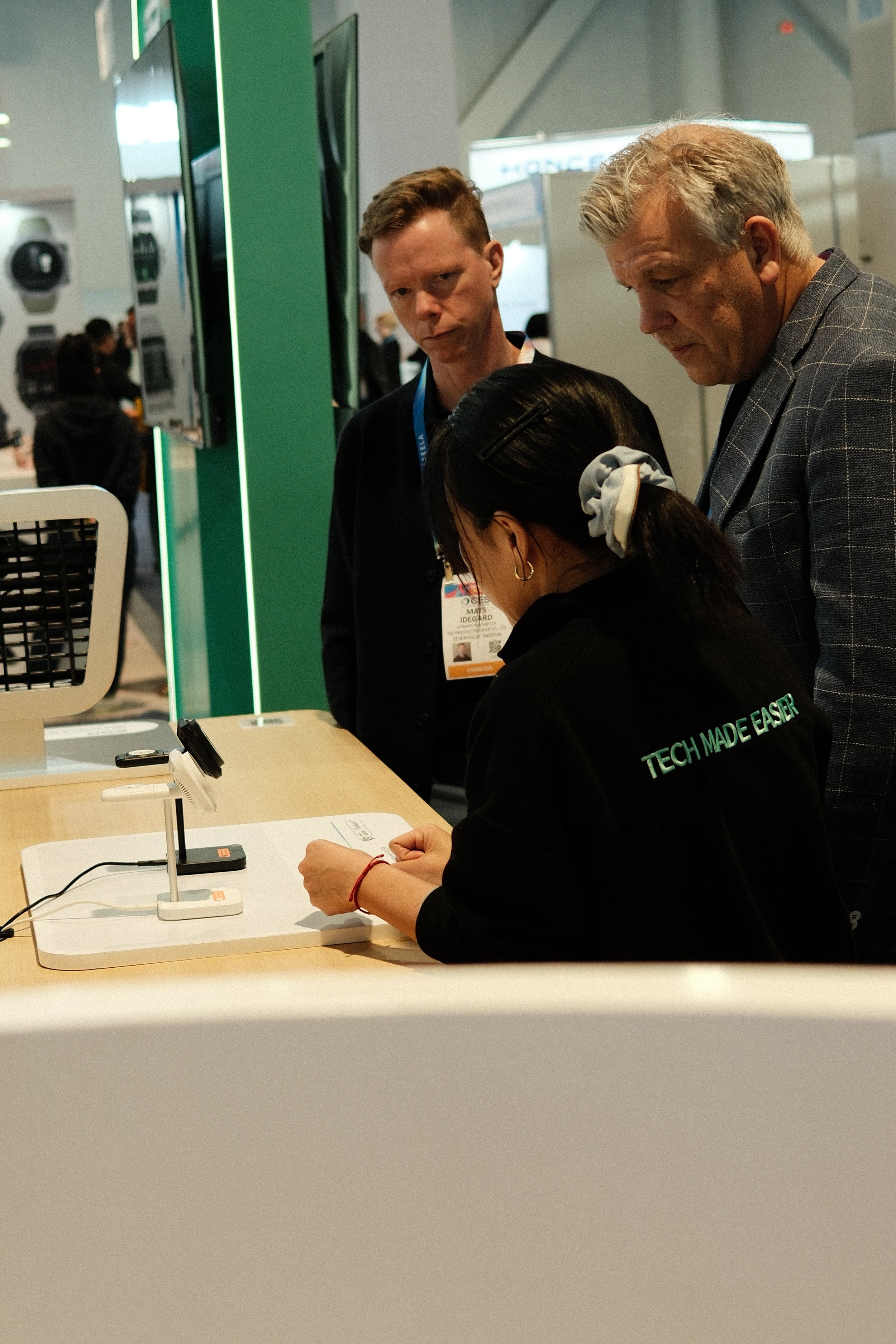 Three people at a booth or display, with one seated person using a device, and two standing individuals observing. The setting appears to be an exhibition or tech event.
