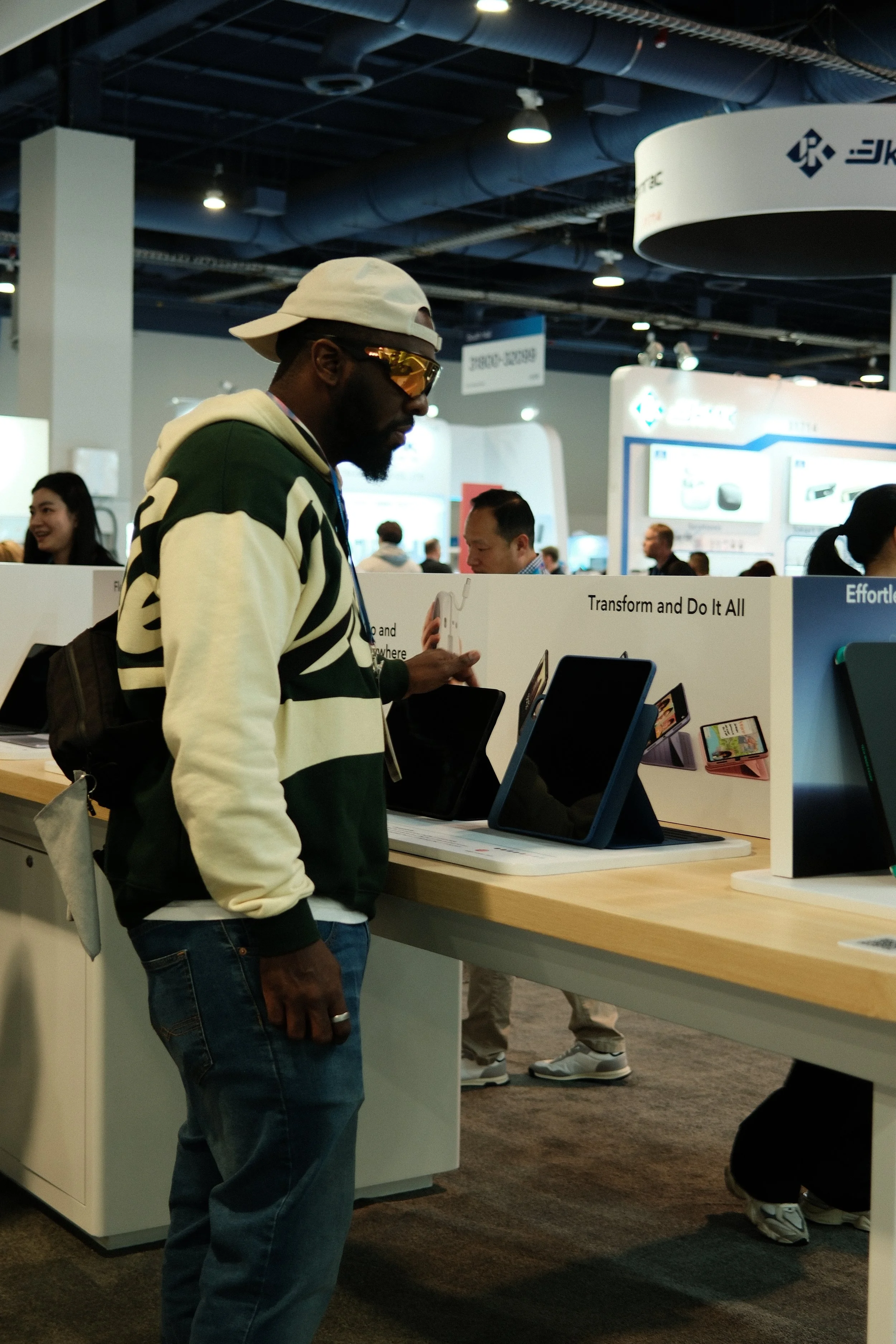 A man wearing a white cap, sunglasses, and a green and white hoodie is looking at tablets on display at a tech store or electronics expo.