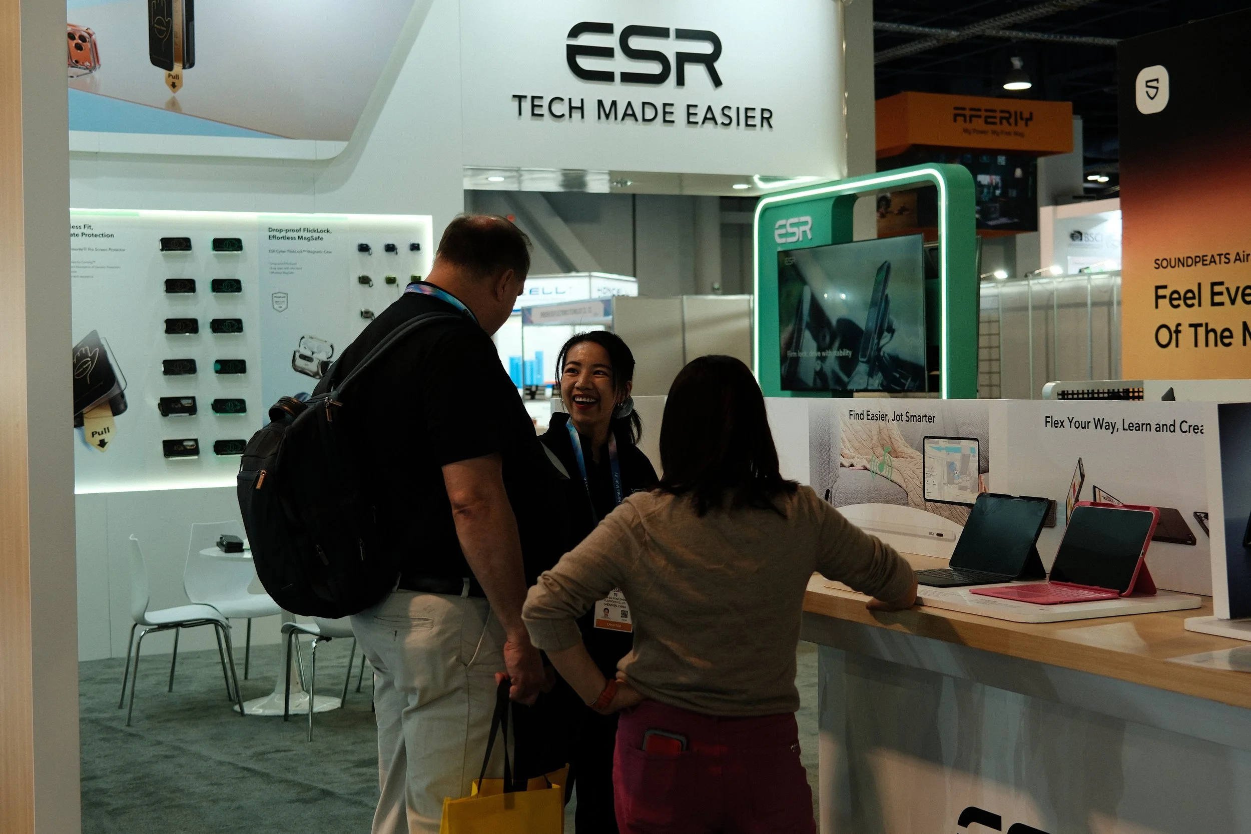 Three people are engaged in conversation at a trade show booth with the ESG branding, featuring electronic gadgets and screens, and a woman smiling.