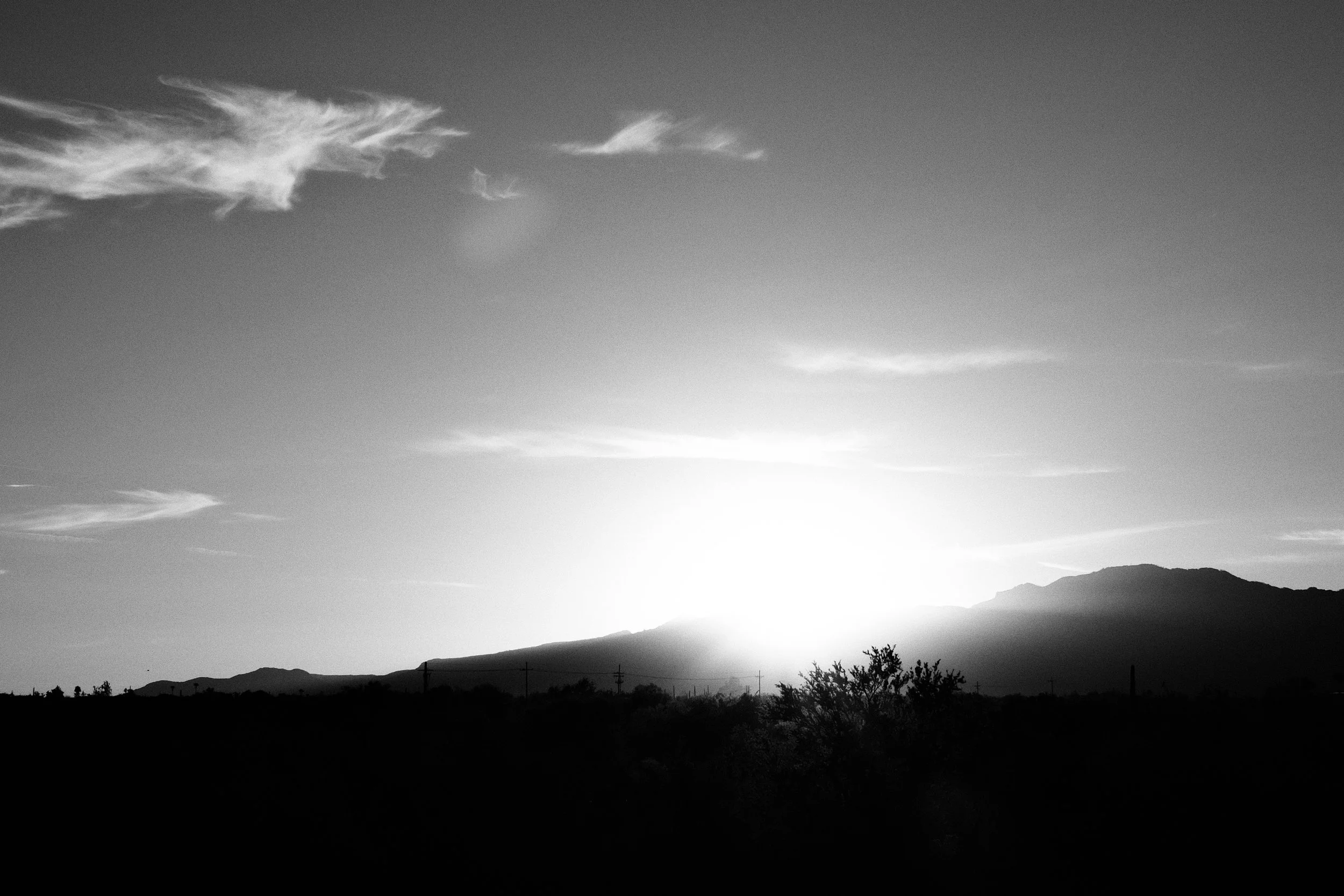 Black and white photograph of a sunset or sunrise over a mountain range with scattered clouds and a silhouetted desert landscape in the foreground.