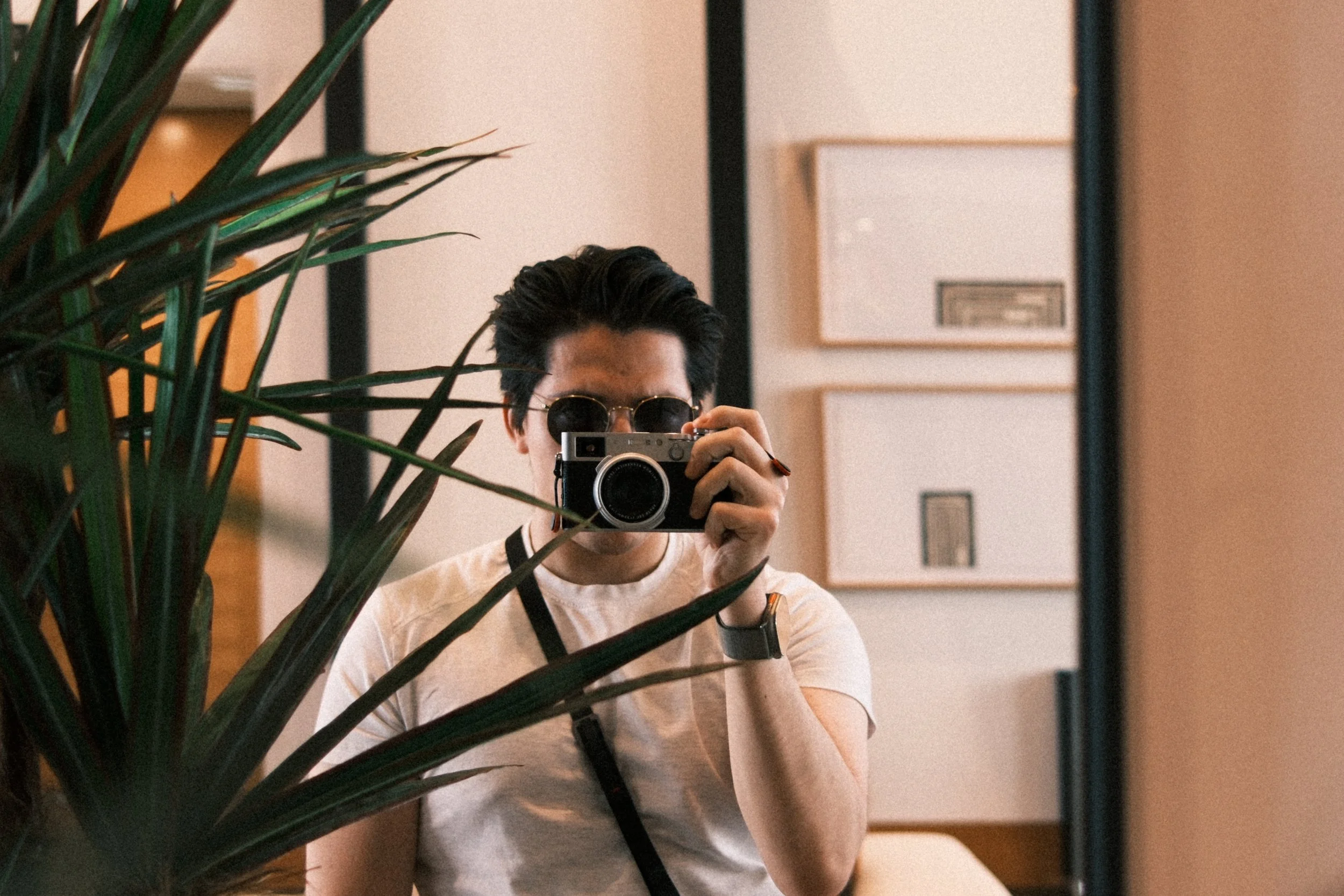 A man taking a mirror selfie with a camera, wearing sunglasses, a white t-shirt, and a smartwatch on his left wrist.