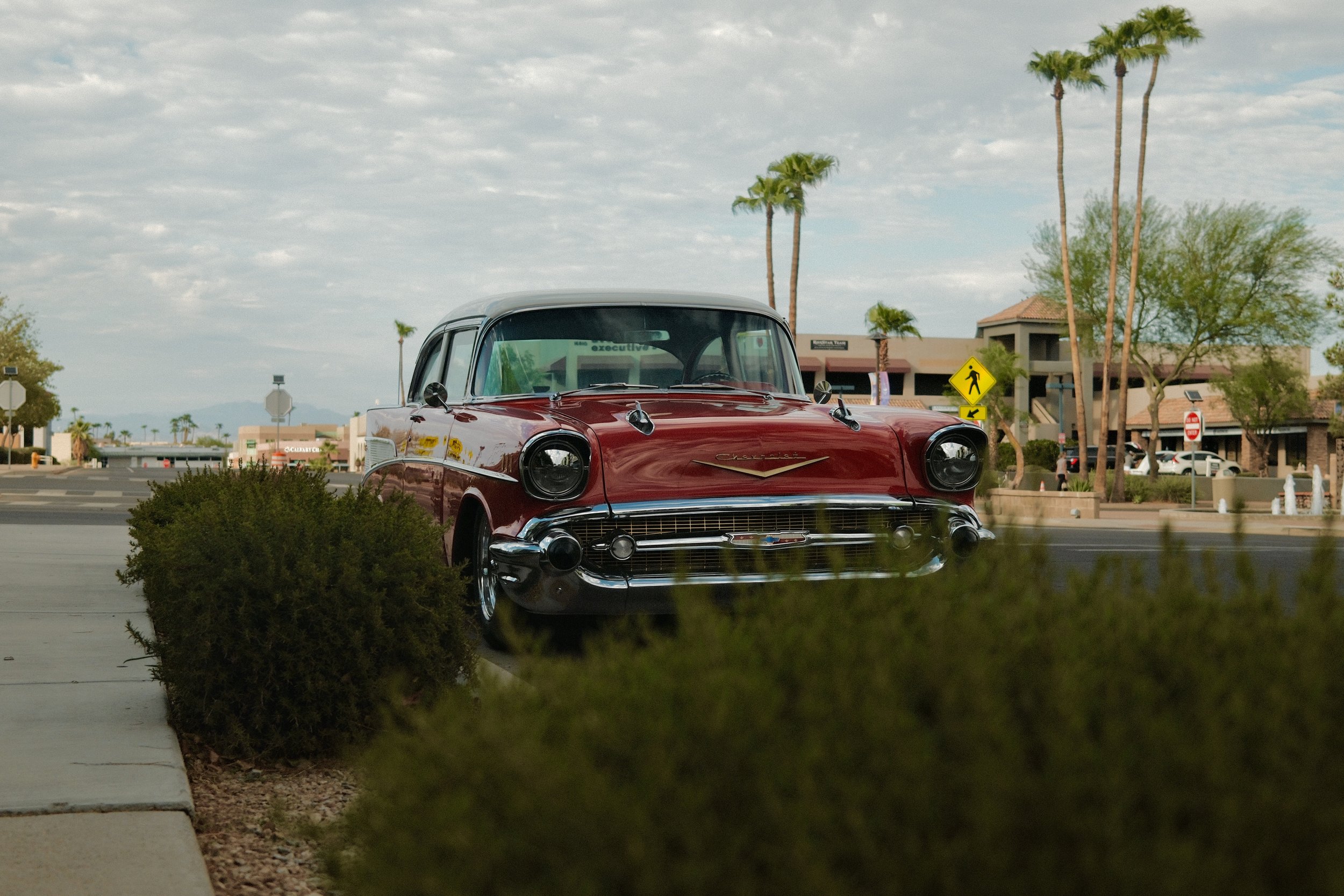 A vintage red Chevrolet car parked on a sidewalk in a suburban shopping area with palm trees.
