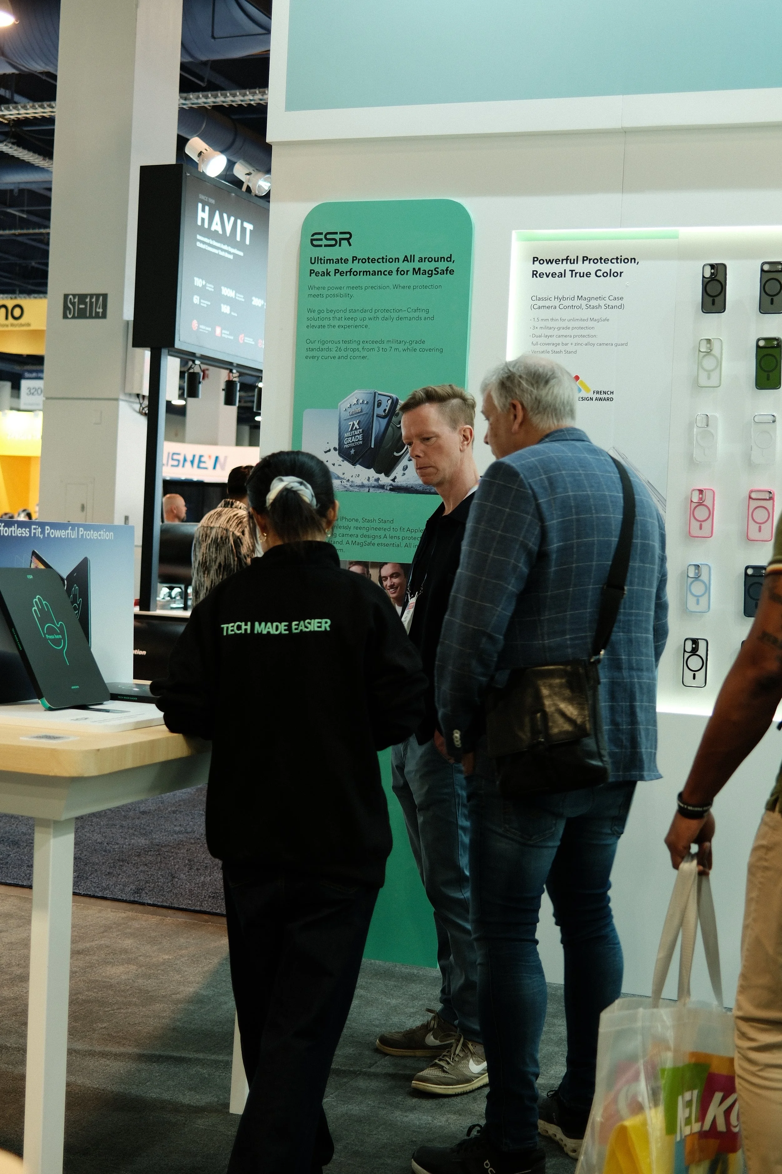 Two men and a woman engaged in conversation at a trade show booth, with informational posters about tech products and protective cases displayed behind them.