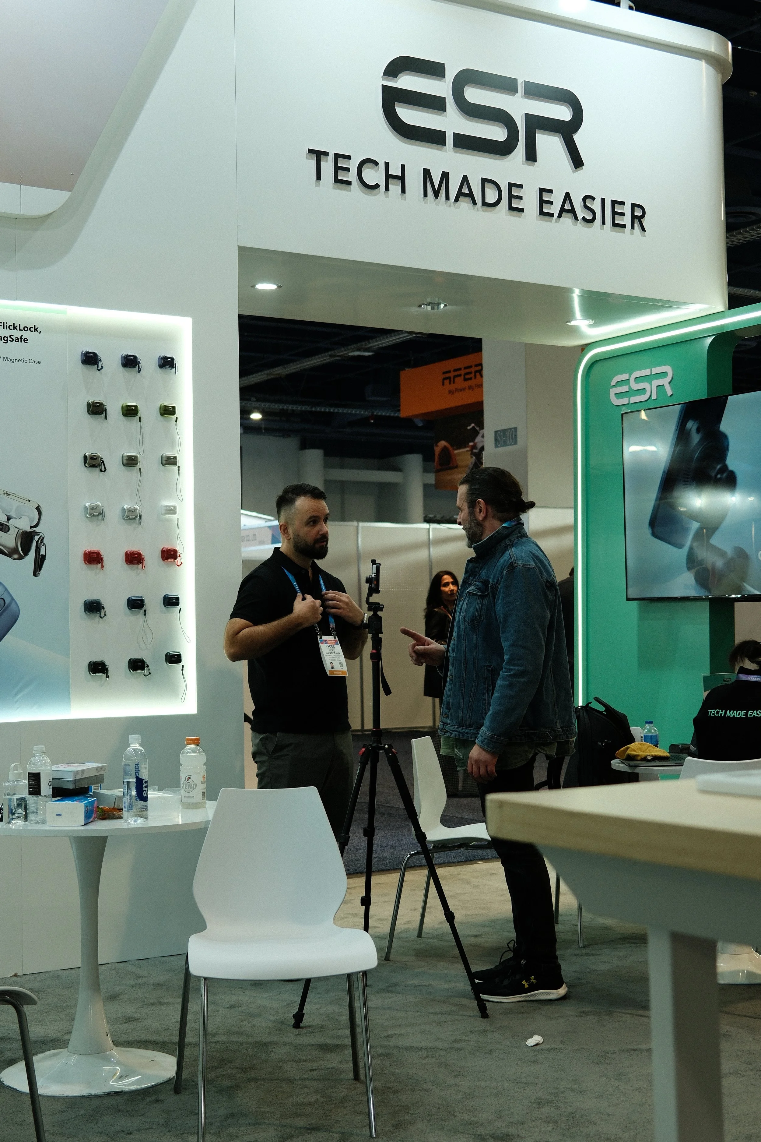 Two men are engaged in conversation inside a technology booth at a trade show. The booth displays the brand 'ESR' with the slogan 'Tech Made Easier.' Various tech accessories are on a panel to the left, and there is a large screen displaying product 
