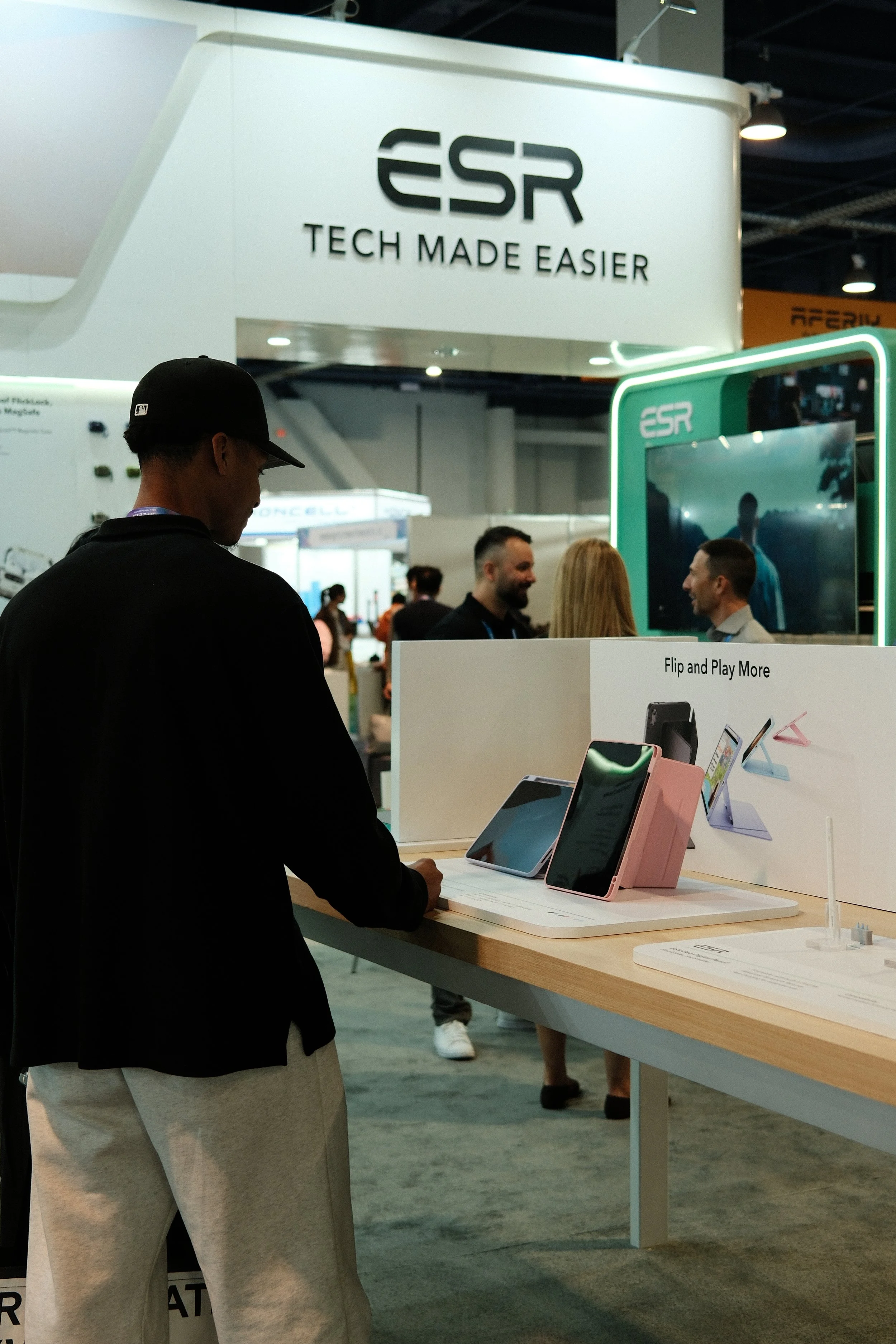 A person wearing a black cap and black jacket is examining electronics at a trade show booth, with multiple smartphones on display and a sign that says "Flip and Play More." In the background, there is a large signage for "ESR" with the tagline "Tech