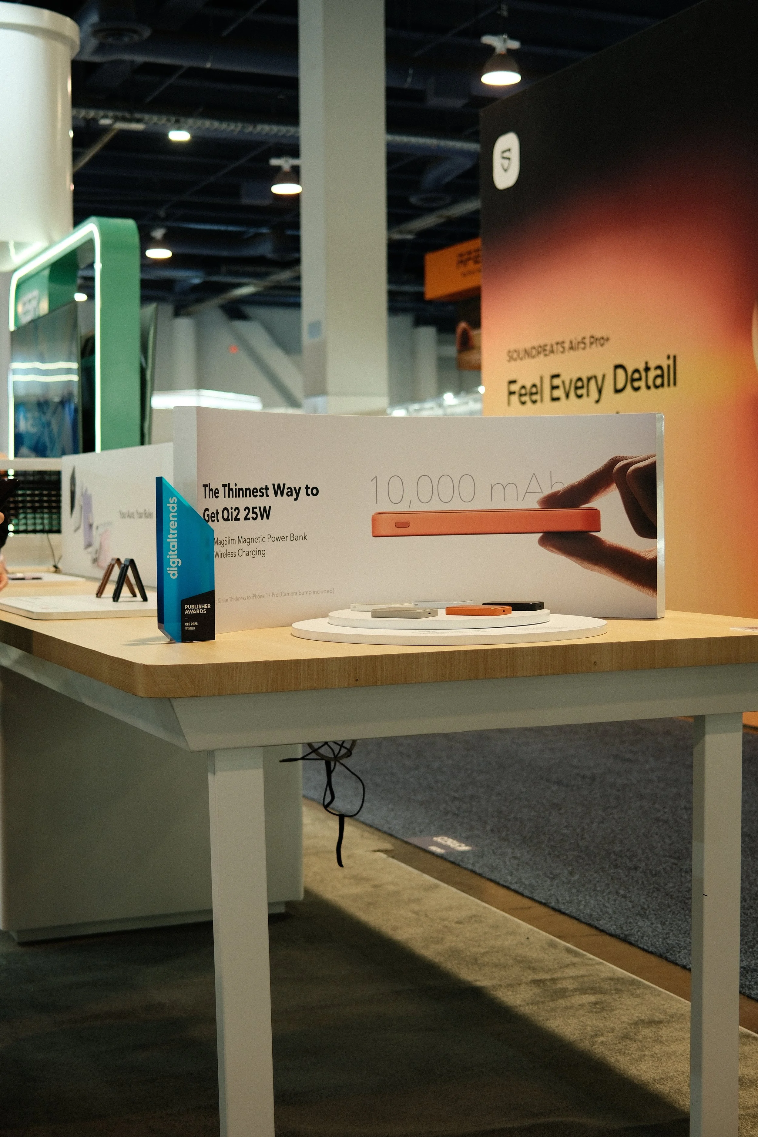 A display table at a technology exhibition showcases a wireless power bank with a 10,000 mAh capacity, labeled as "The Thinnest Way to Get Qi2 25W." The setup includes promotional signage and a few small electronic devices.