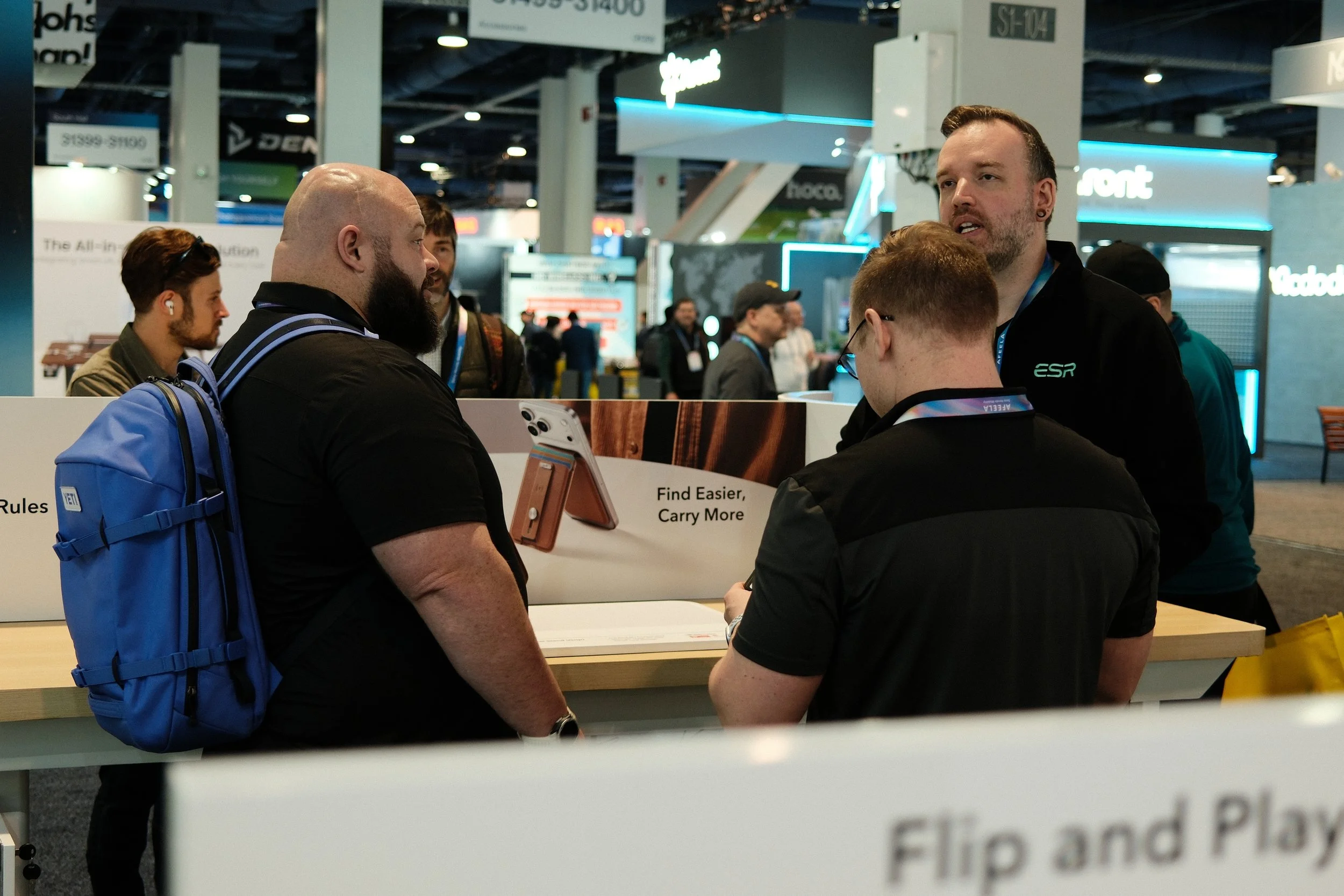 Group of people at a technology exhibition booth, engaging in discussions about a product with a sign that reads 'Find Easier, Carry More' and electronic gadgets on display.
