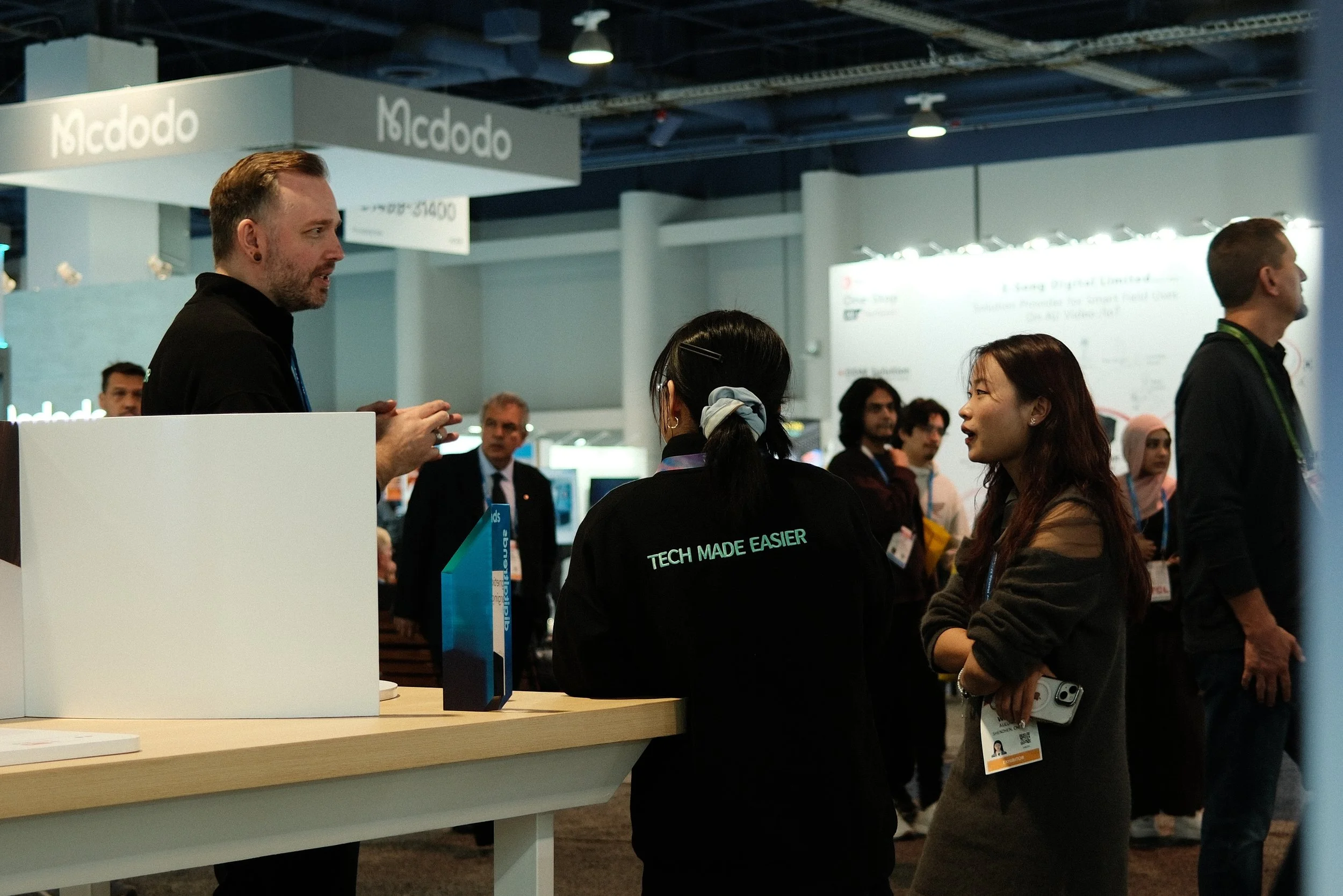 People attending a technology trade show or conference, engaging in conversations, with booth signage displaying 'McDodo' and informational displays in the background.