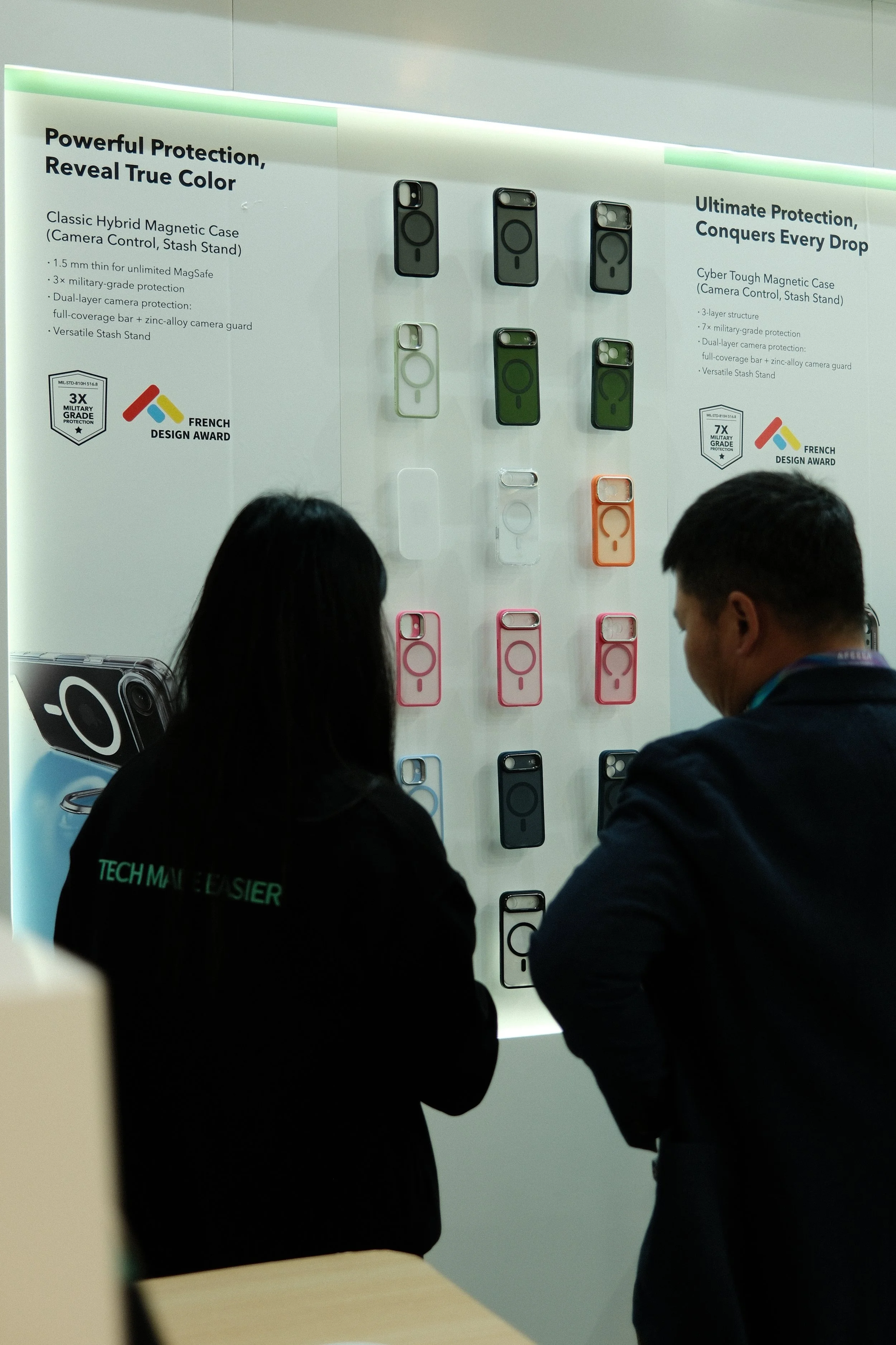 Two people viewing a display of various protective phone cases at an exhibition, with informational signage about the features and design awards of the cases.