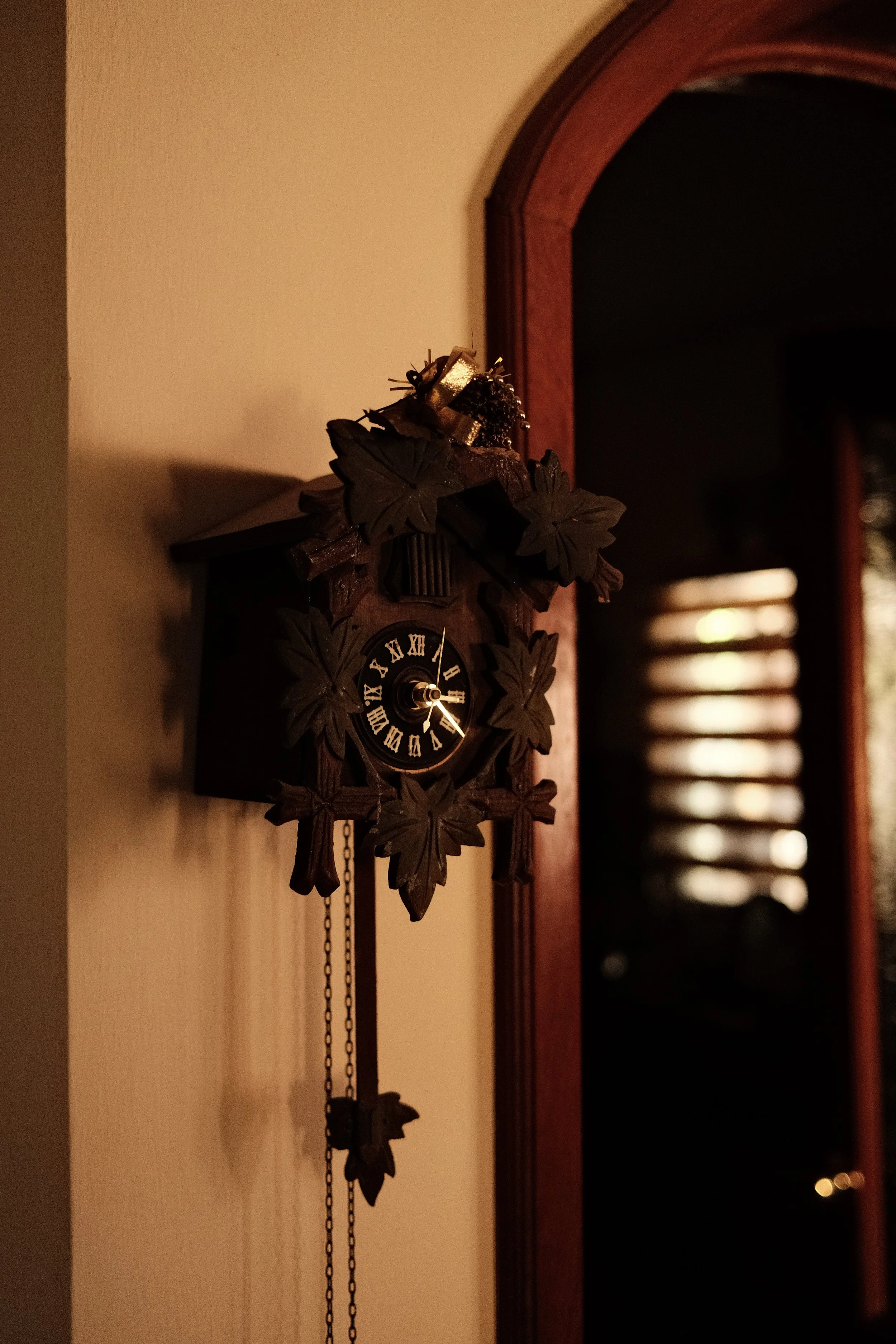 A traditional wooden cuckoo clock with leaf and bird carvings hanging on a beige wall in a dimly lit room, with a doorway with reddish-brown trim in the background.