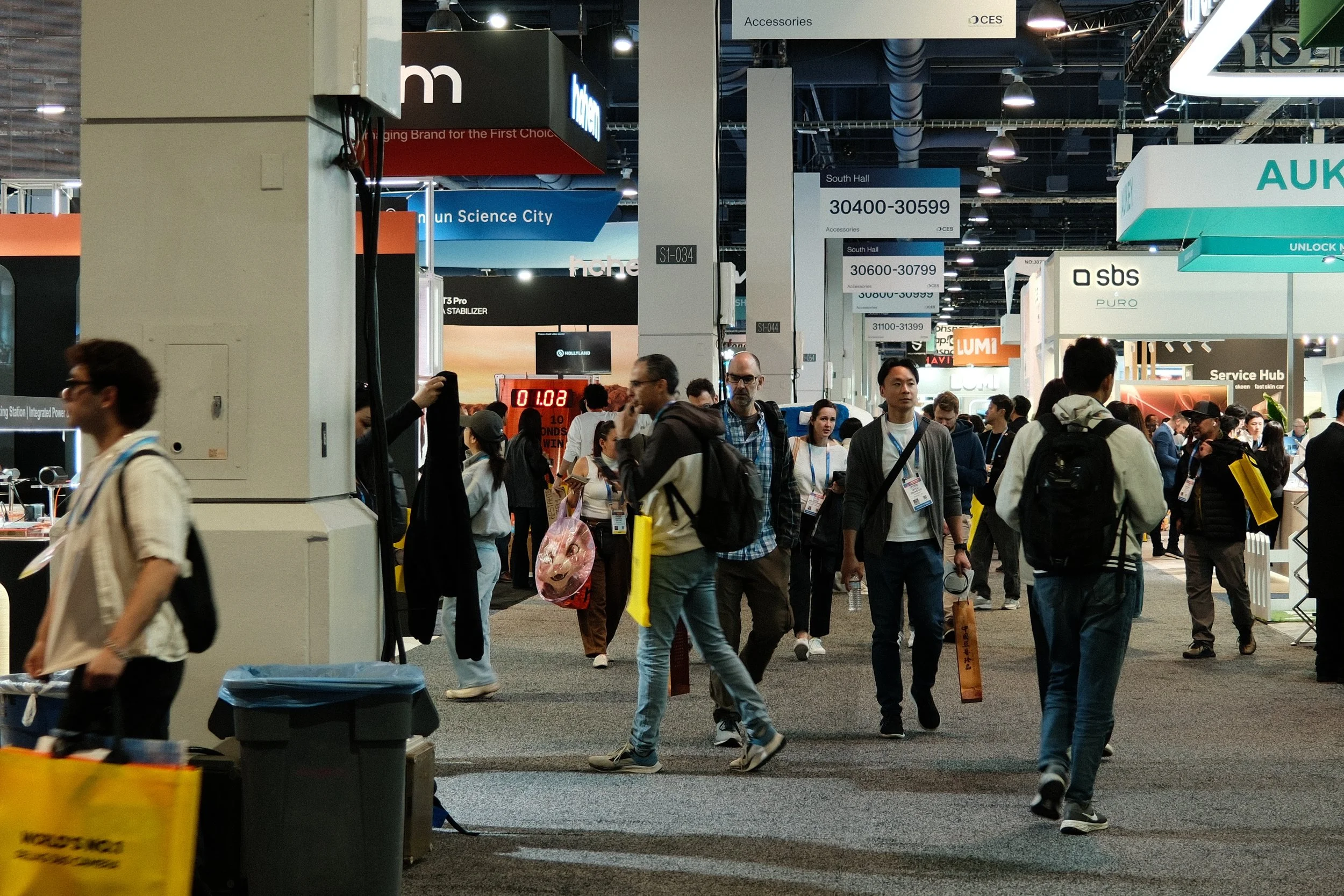 A busy scene at a trade show or convention with many people walking and looking at booths under signage indicating booth numbers and company names.
