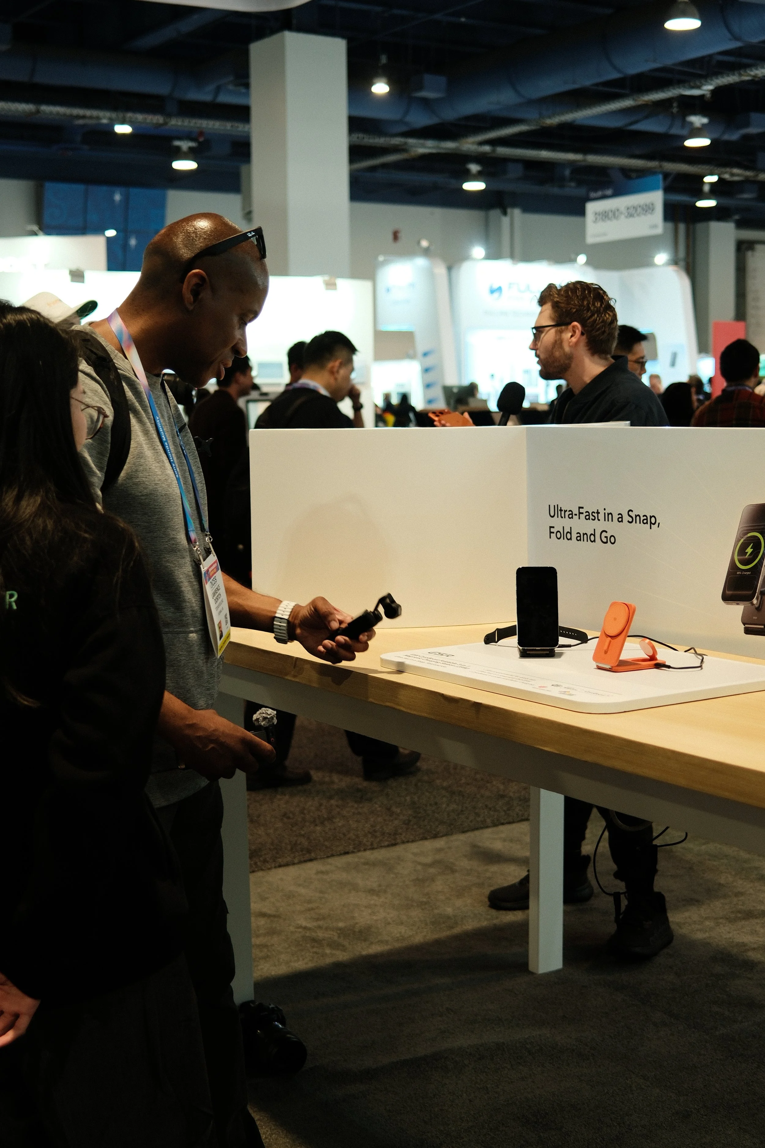 A person at a trade show or technology event demonstrating wirelessly charging a smartphone placed on a small orange stand, with a sign that reads 'Ultra-Fast in a Snap, Fold and Go.'