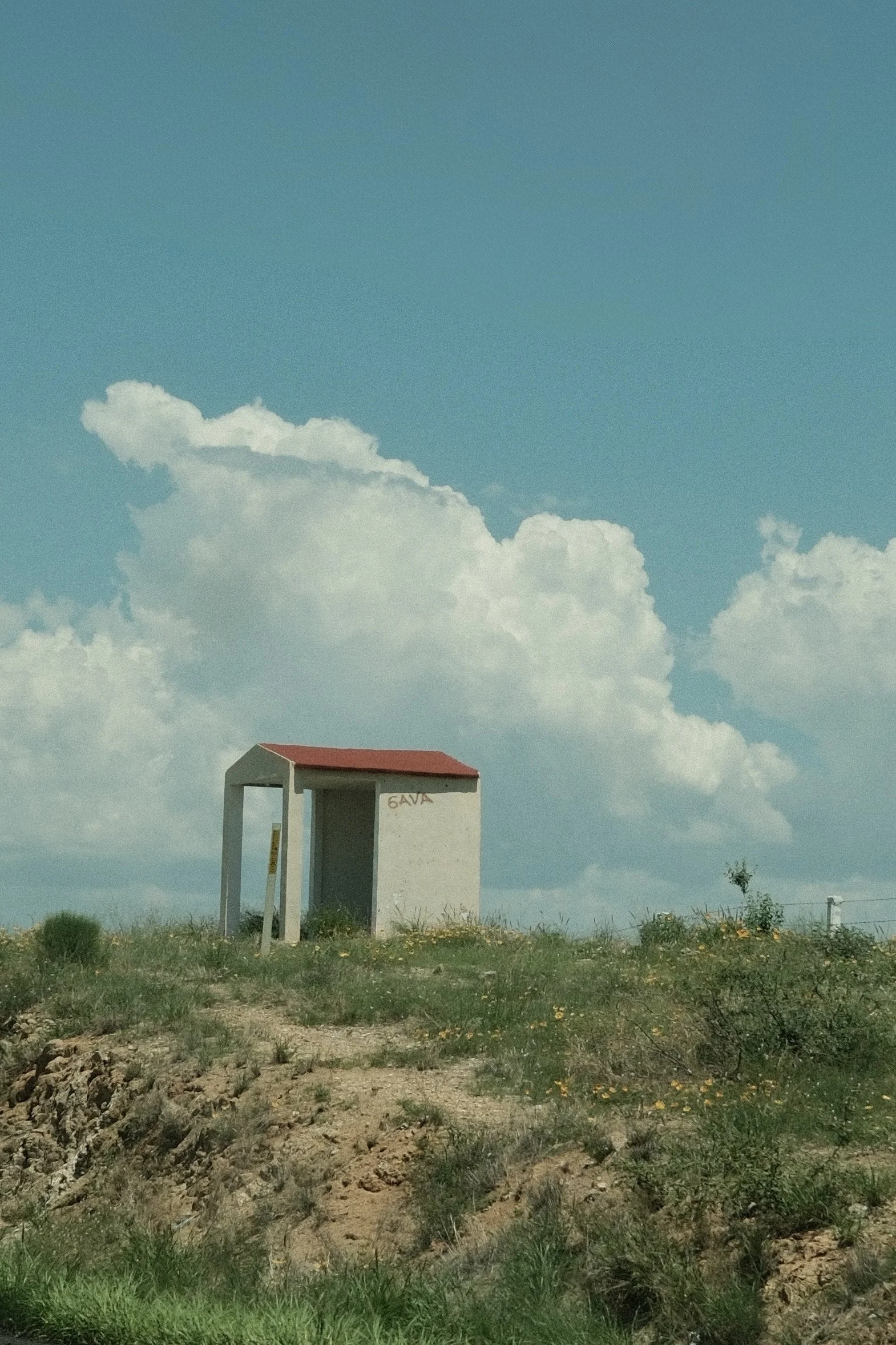 A small, simple structure with a red roof and white walls situated on a grassy hill under a blue sky with fluffy clouds.