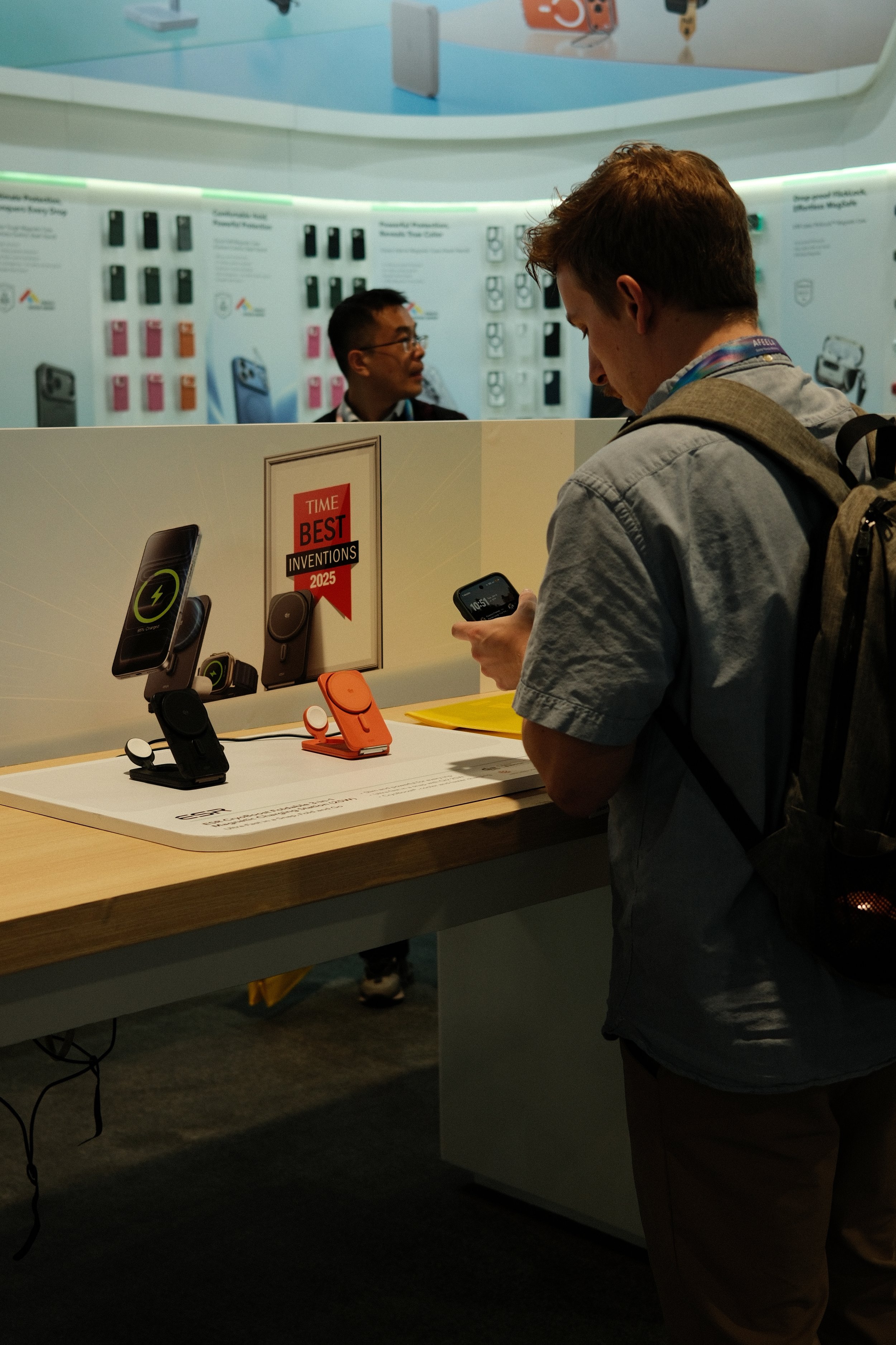 A man with a backpack looking at a smartphone at a display table showcasing wireless phone chargers, with a sign indicating they are Time Best Inventions of 2025.