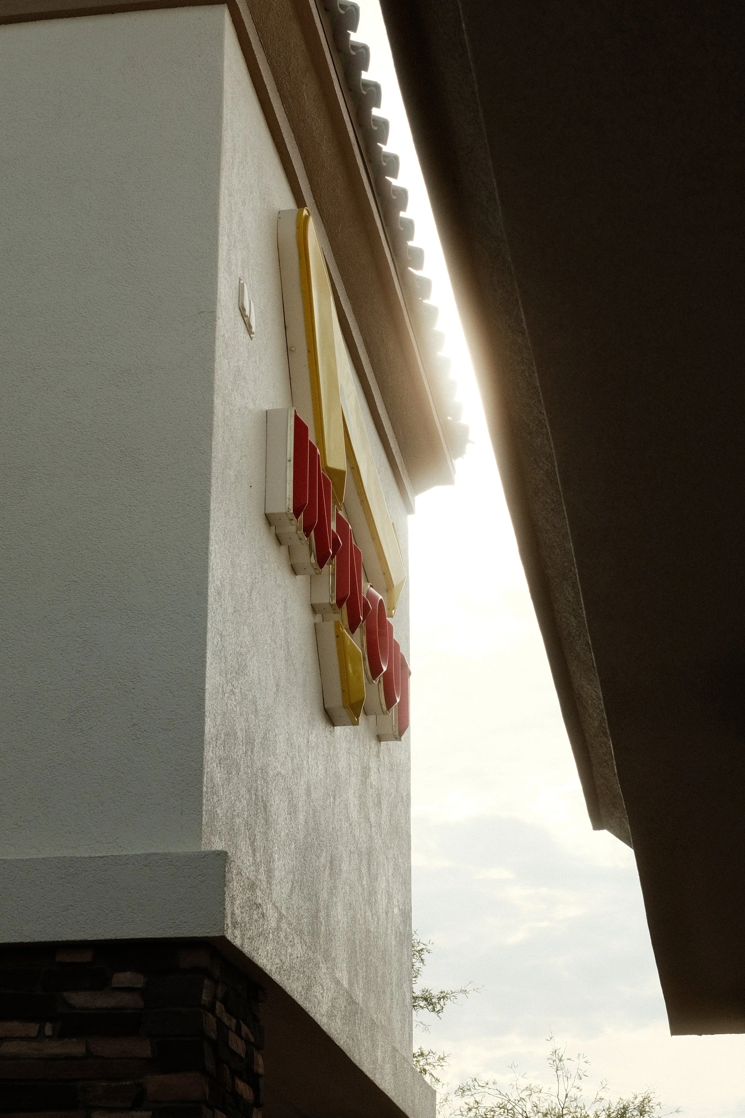 Close-up of a retro style sign with yellow and red elements, mounted on a beige wall of a building with a dark overhang and a cloudy sky in the background.