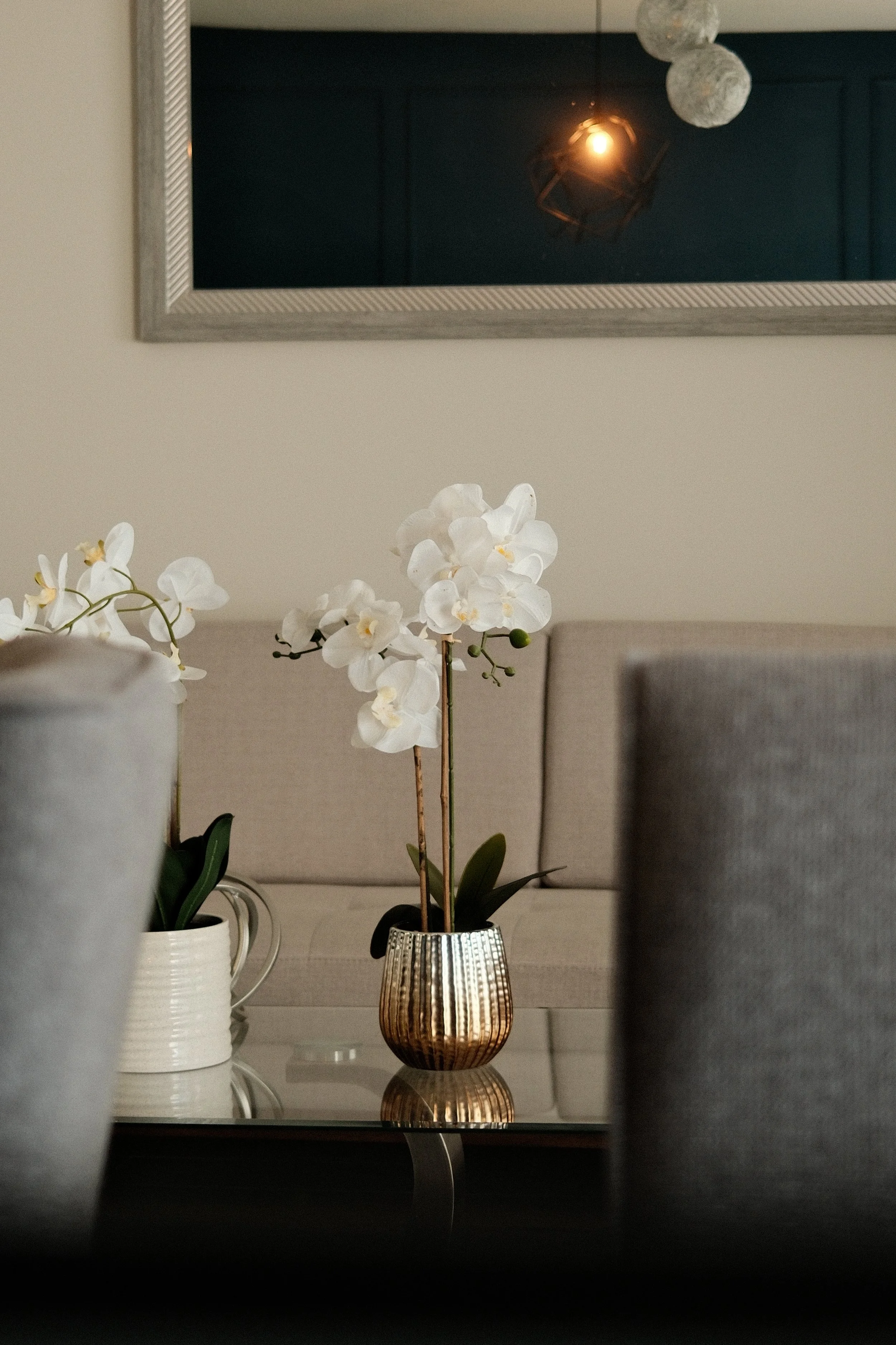 A white orchid in a metallic gold vase on a dark dining table, with upholstered chairs and a mirror reflecting a hanging light fixture in the background.