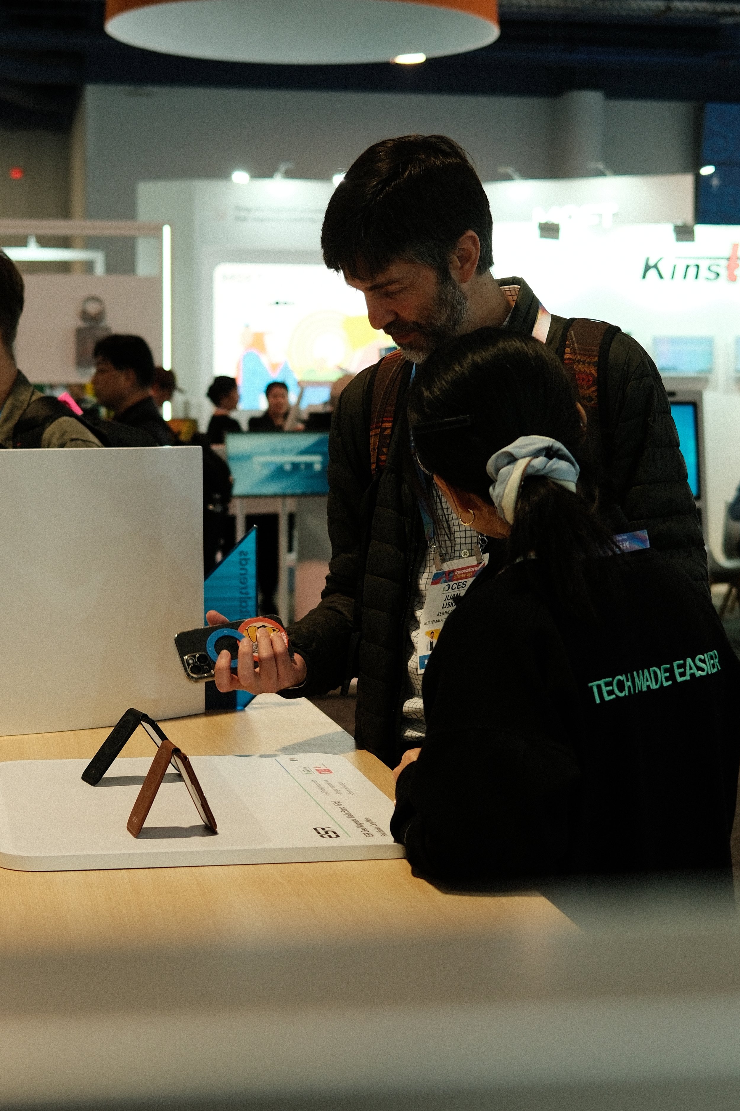 A man and a woman are looking at a smartphone together at a technology trade show or exhibition. The woman is wearing a black jacket with the phrase 'TECH MADE EASIER' on the back.