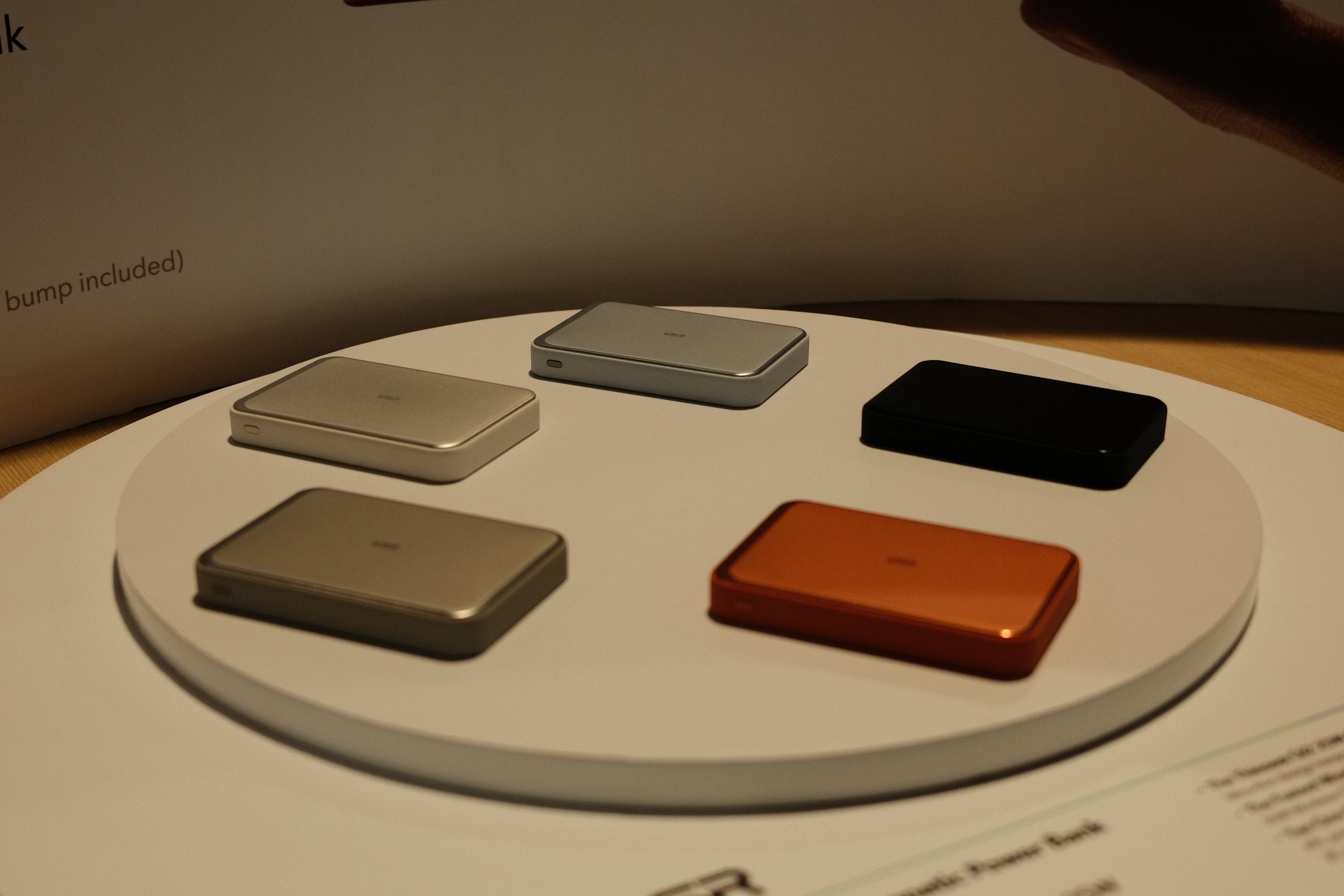 Six closed rectangular electronic devices in different colors (gold, silver, black, copper) are displayed on a round white surface, with a brochure in the background.