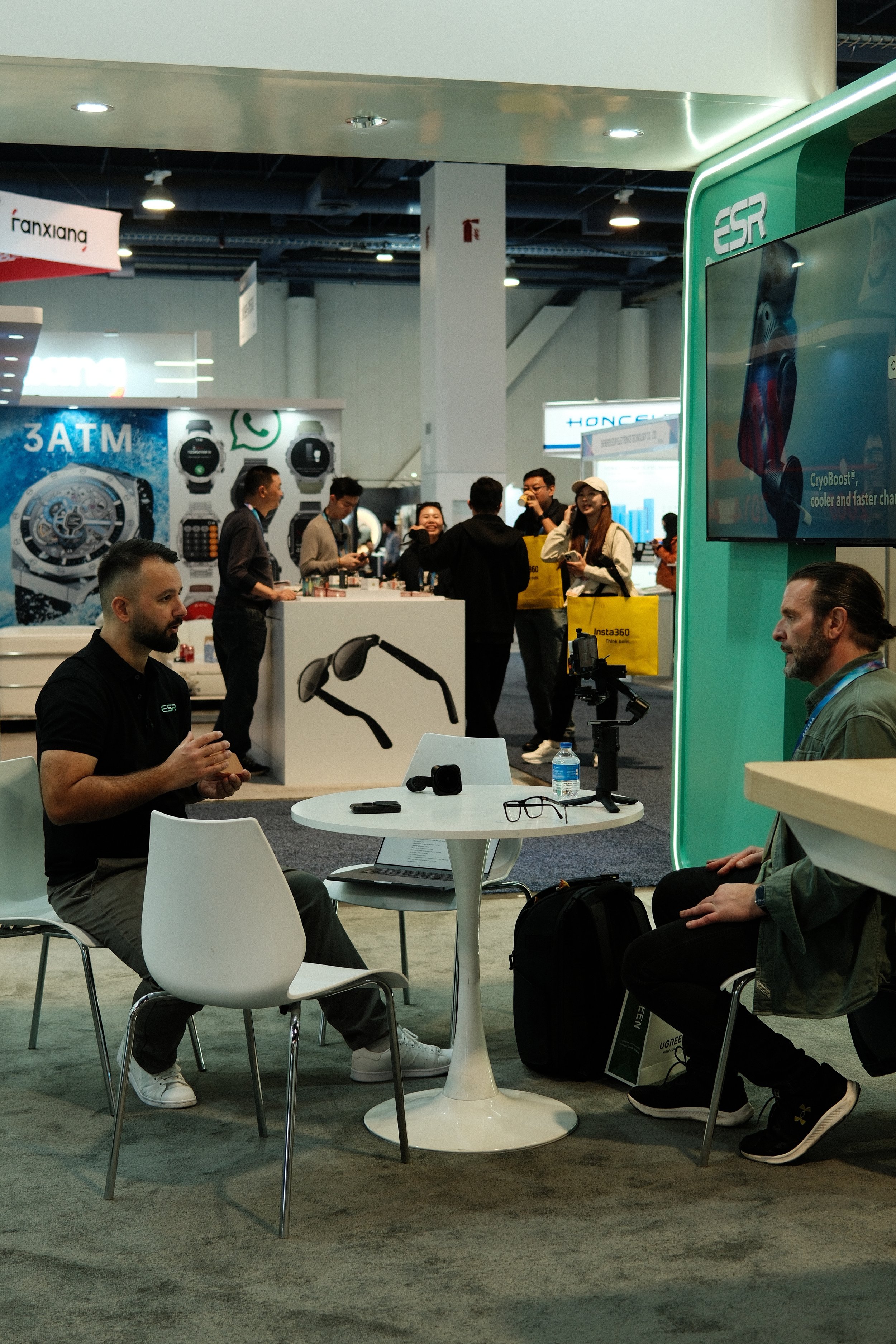 Two men seated at a small round table having a conversation at a tech expo, with electronic gadgets, including a laptop and glasses, on the table, and a large screen and promotional materials in the background.