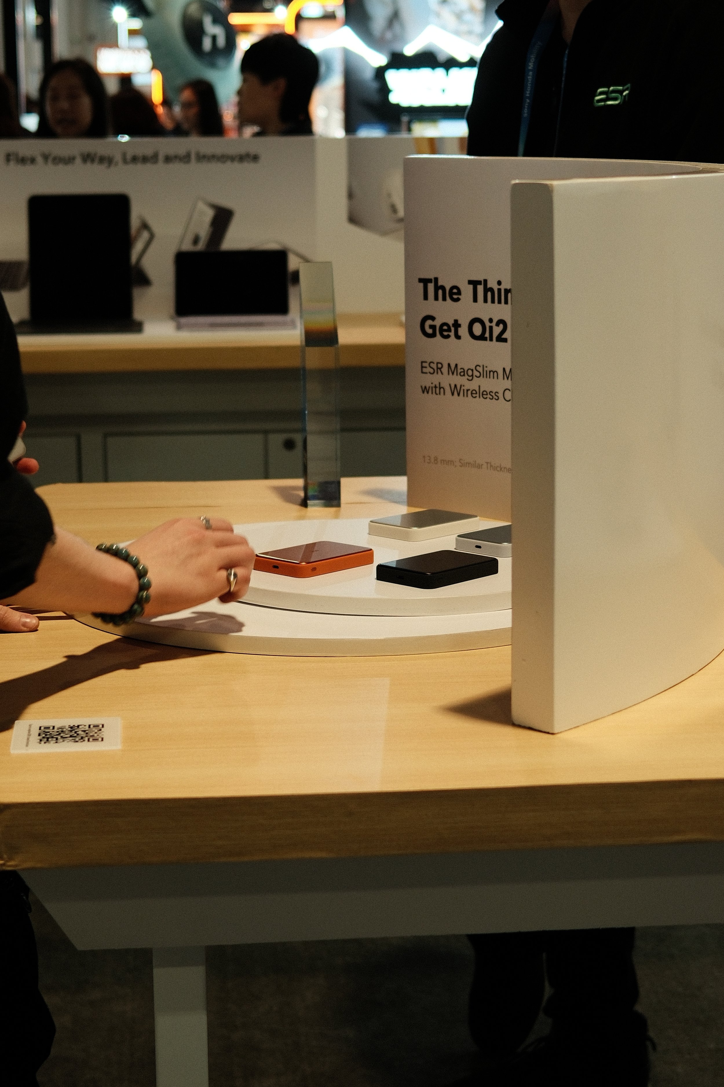 A person browsing smartphones displayed on a round white table at a tech store or exhibition. Multiple phones in various colors are visible with a sign promoting the Qi2 wireless charging standard.