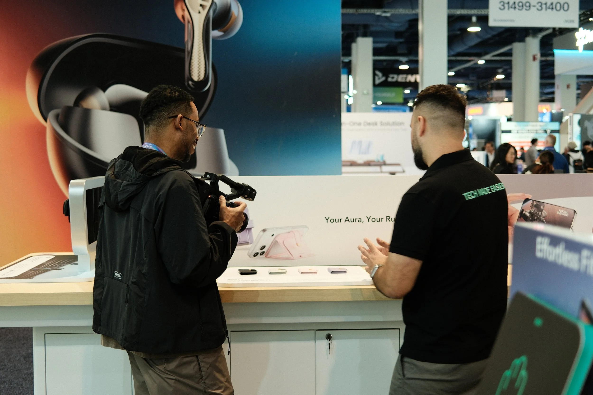 Two men in conversation at an electronics booth, one holding a camera, in a busy trade show or tech expo setting.