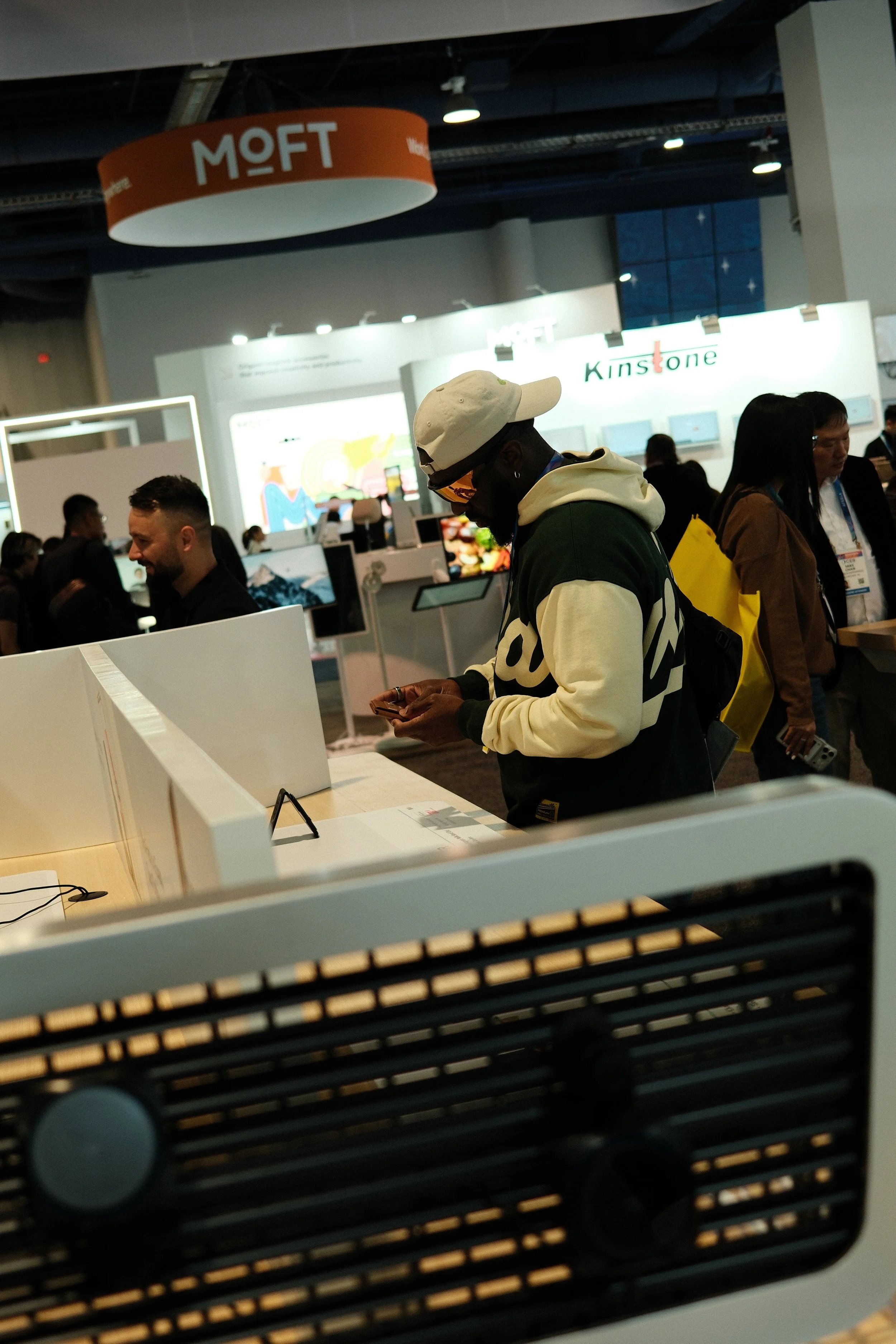 A man wearing a beige cap, sunglasses, and a hoodie with a backpack, looking at his phone at a busy trade show or exhibition.
