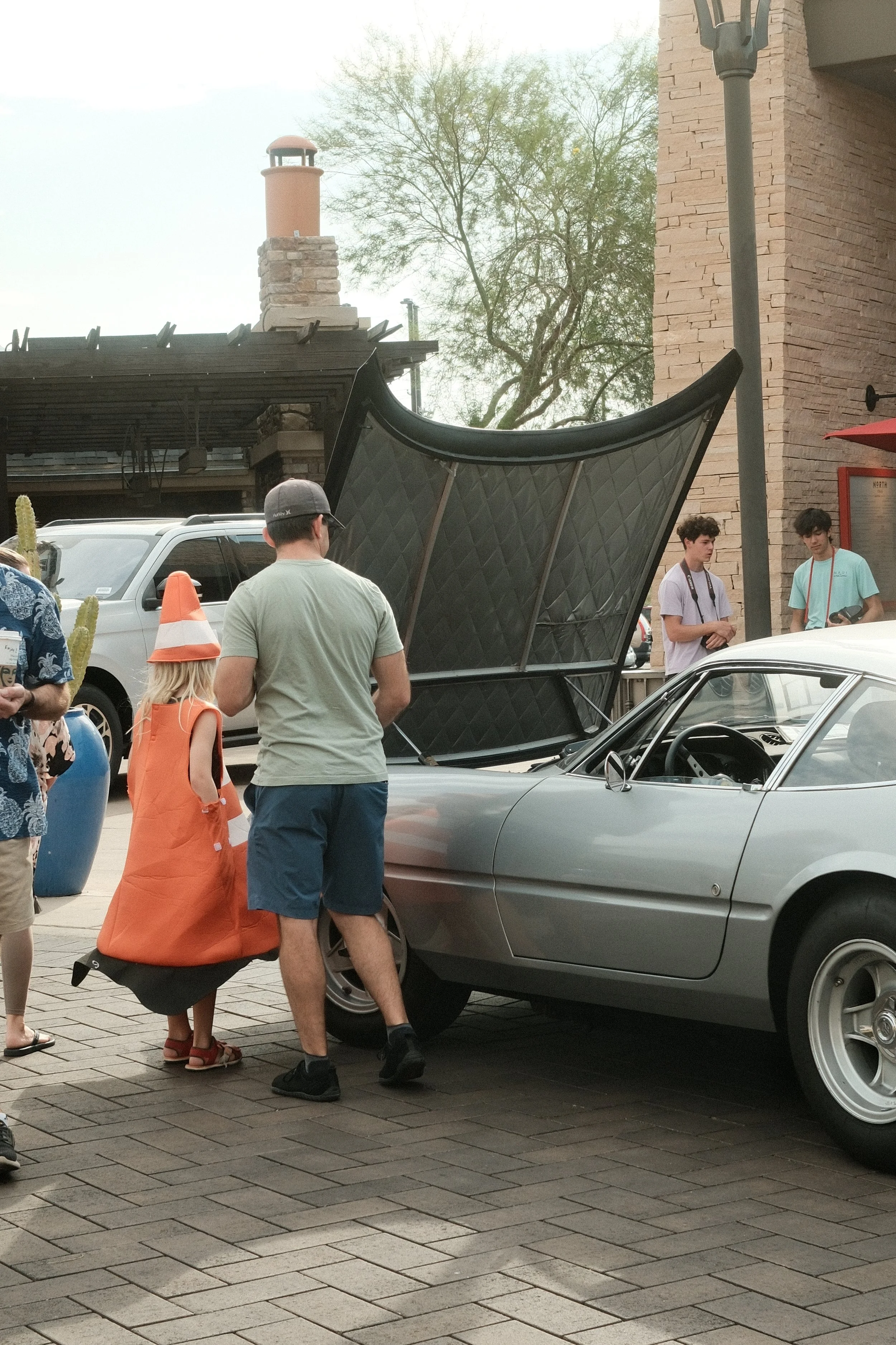 People standing and walking near a silver vintage sports car with its hood open, at an outdoor car show.
