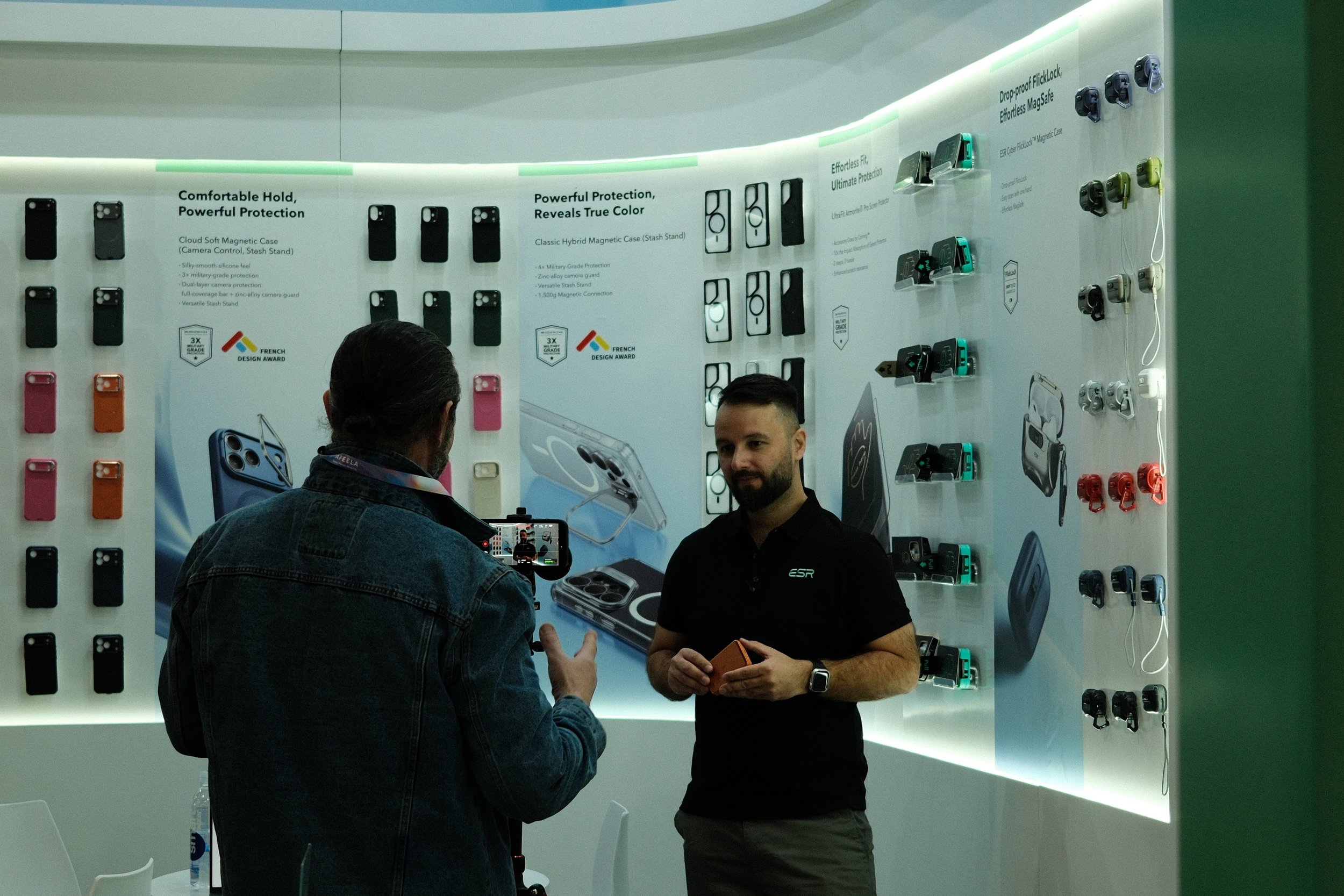 Two men at a tech display booth showcasing phone accessories, with one man recording or taking a photo of the other.