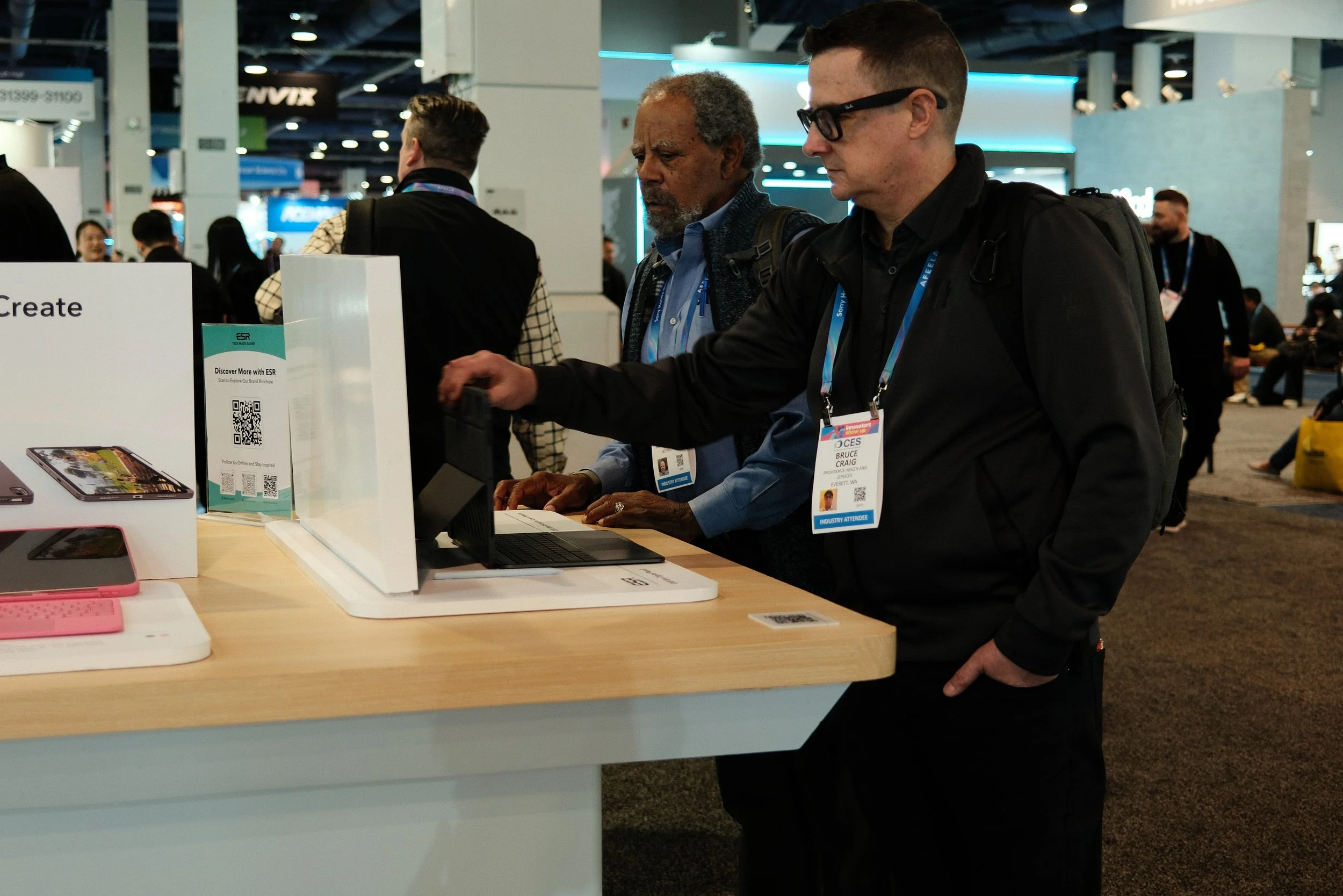 Two men with conference badges interact with a laptop at a trade show booth, while other attendees walk in the background.