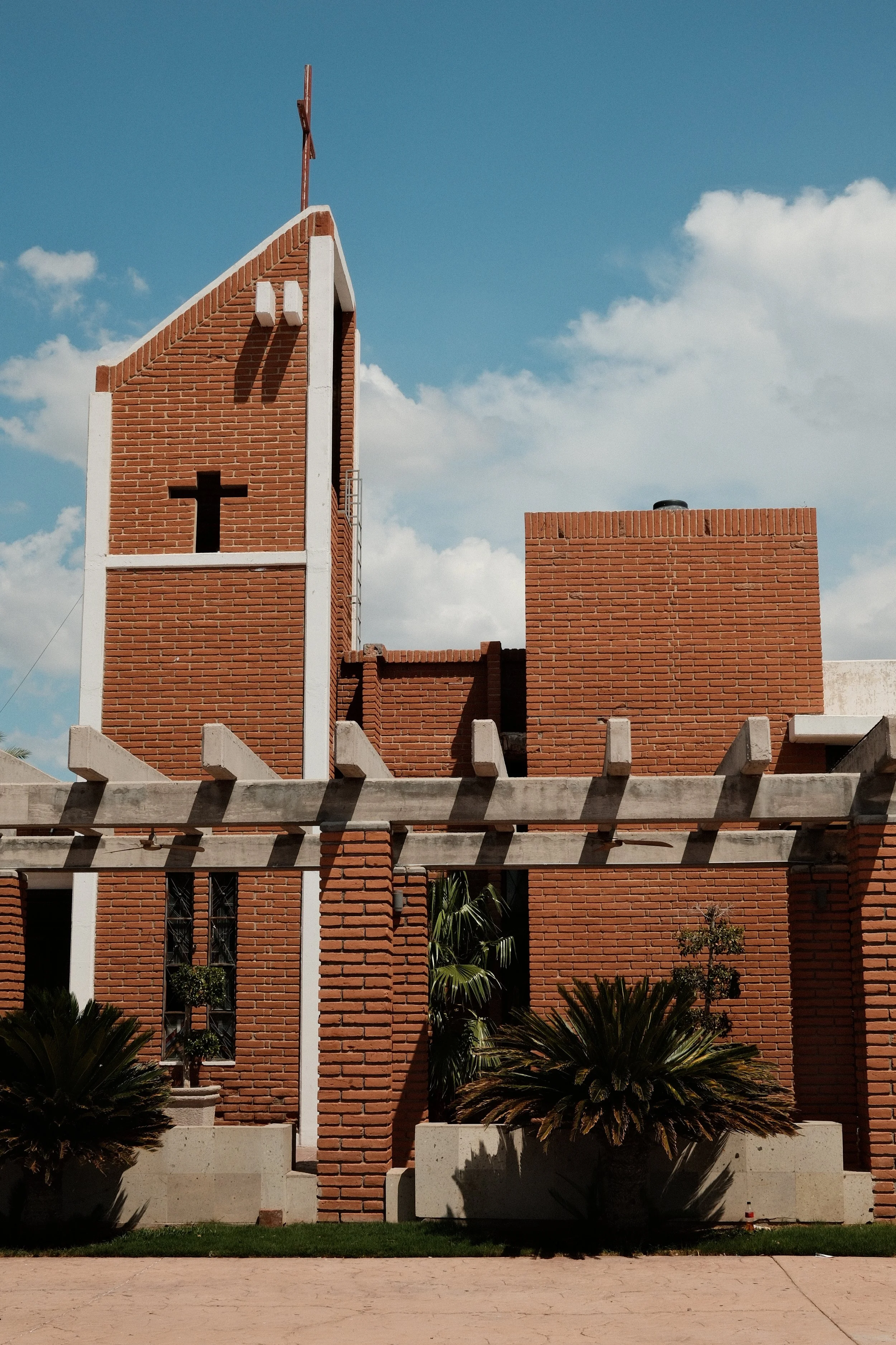 Brick church with a cross on top, white accents, a large cross-shaped window, plants in front, and a blue sky with clouds.
