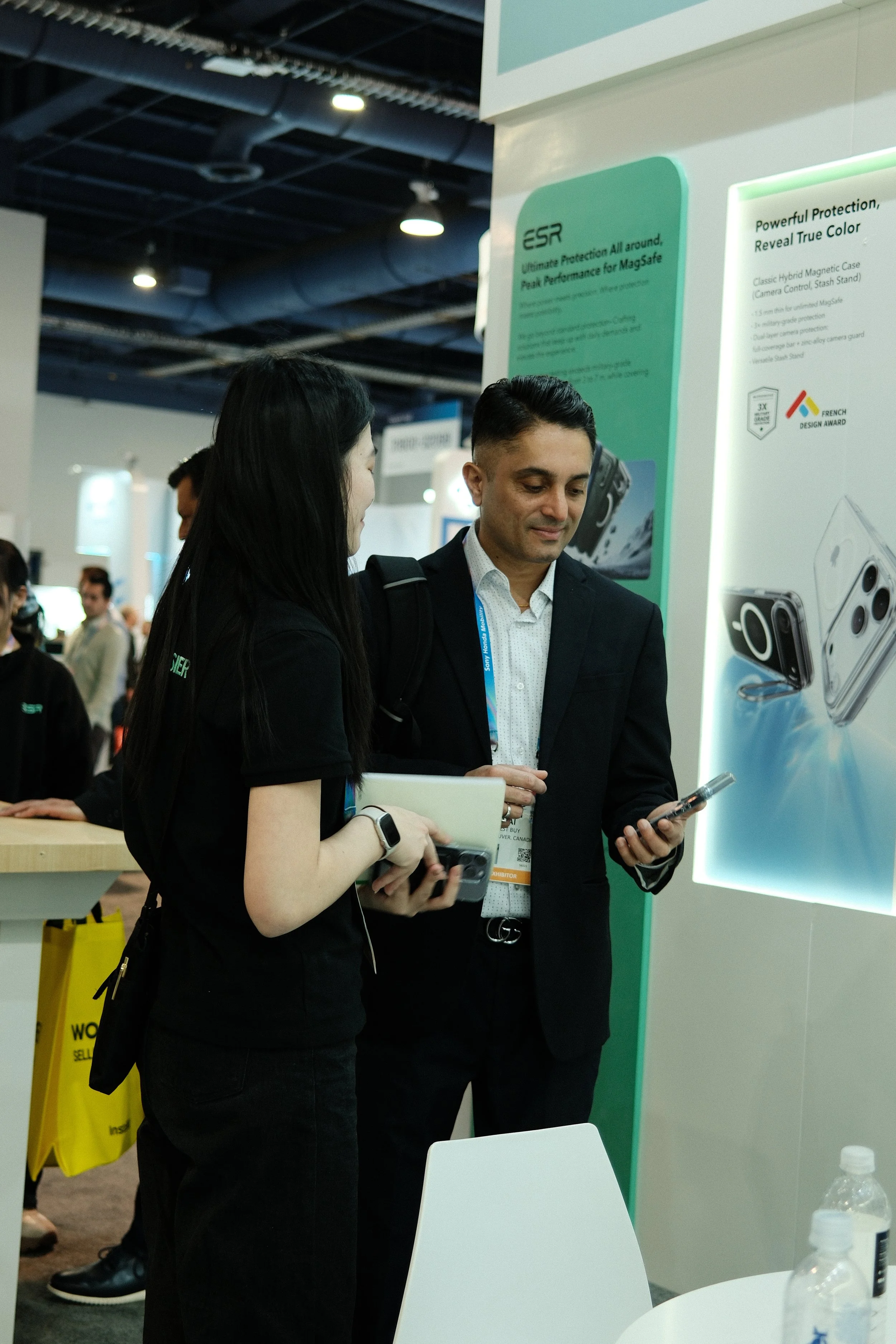 Two people look at their phones in front of a display with tech product advertisements at an exhibition or trade show.